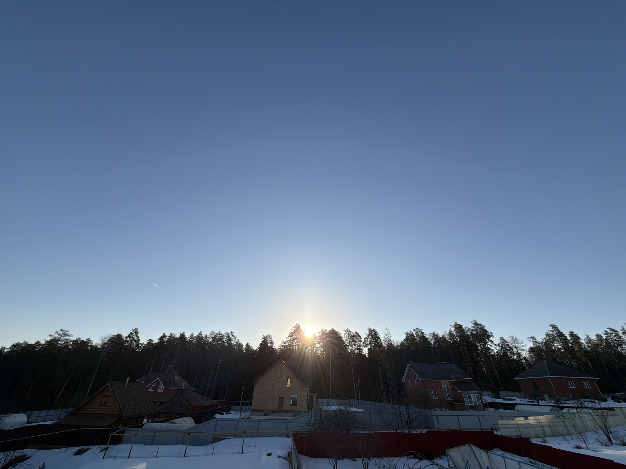 The image shows a clear, light blue sky above a residential area. The sun is setting or rising behind a line of tall, dark green trees, creating a sunburst effect. Below the trees are several houses, varying in style, with snow covering the ground. Fences are visible between the properties. The location is Chaikovsky, Russia.