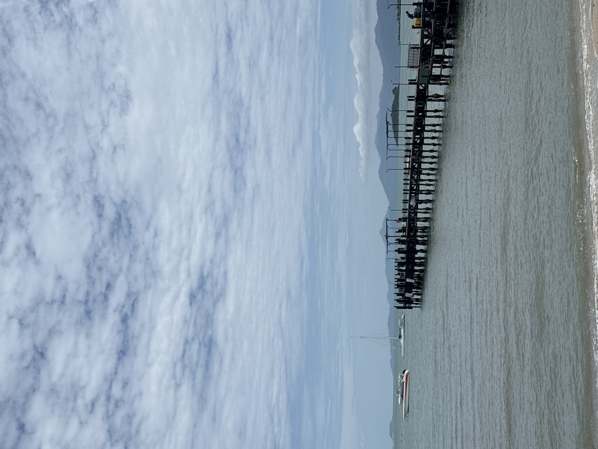The image depicts a wide shot of a pier on the coast of Ko Samui District, Thailand, under a cloudy sky. The pier, constructed of dark wood and supported by numerous pilings, extends from the right side of the frame into the ocean. The water is a muted green, with small waves lapping at the sandy shore in the foreground. Several boats are visible on the water, including a white speedboat with red accents on the left and a white sailboat further out. In the background, a chain of green mountains forms the horizon. The pier itself is lined with railings and has several structures, including what appears to be a covered area with a person visible. The sky is filled with layered, wispy white clouds against a pale blue backdrop.
