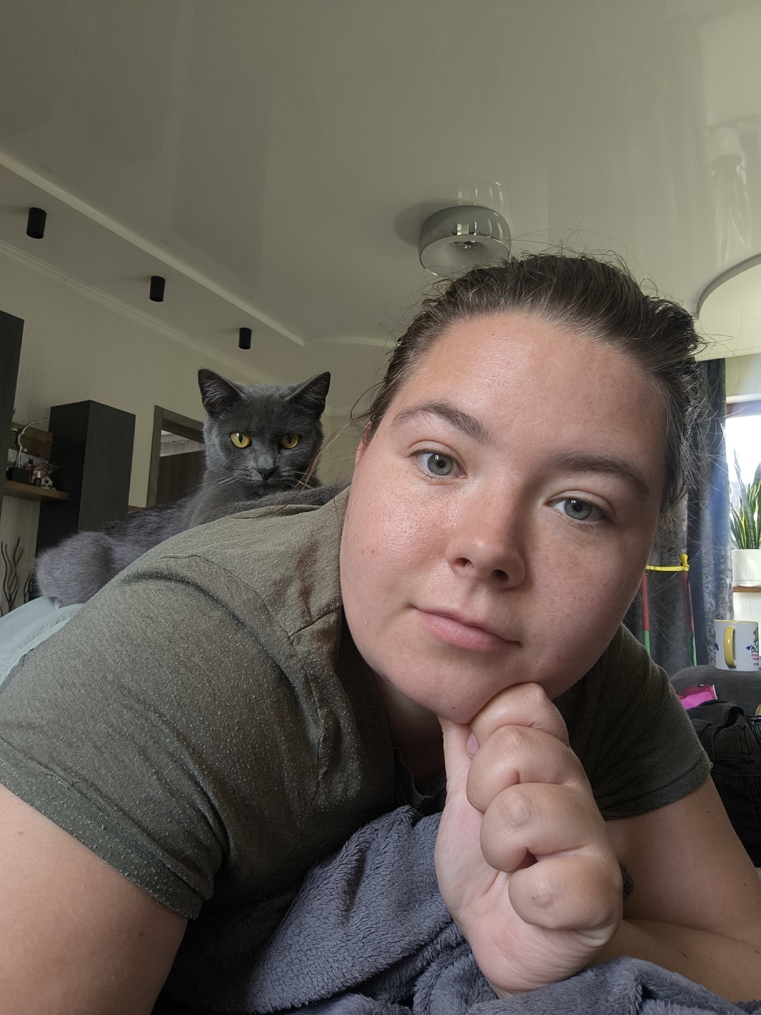 The image shows a close-up selfie of a woman with her hand resting on her chin, looking towards the camera. A gray cat is perched on her shoulder, also looking at the camera. They appear to be indoors, possibly in a bedroom, with a ceiling and lighting fixtures visible in the background. The woman is wearing a dark green t-shirt. The cat has yellow eyes. The setting is not identifiable as being in the city of Kaliningrad, Russia.