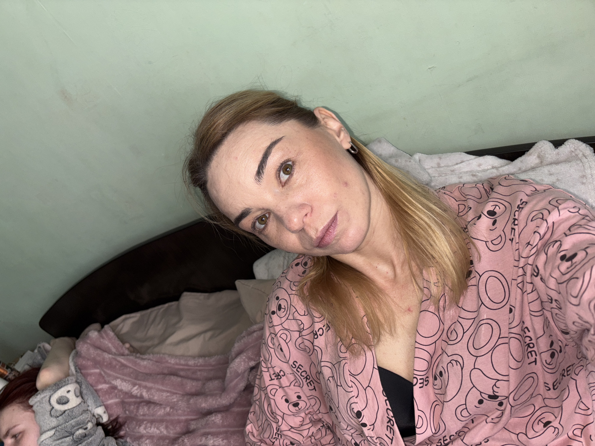 The image shows a medium shot of a woman with blonde hair and light skin. She is lying in a bed, with her head tilted to her left. She is wearing a pink robe with a pattern of teddy bears and the word "SECRET" repeated. A black bra is visible underneath the robe. Her eyes are open and looking towards the camera, and she has a slight smile on her lips.
To the left of the woman, a child is partially visible, also in bed. The child is wearing grey pajamas with a teddy bear pattern and a pink blanket is covering them. The bed is set against a dark headboard, and the wall behind the bed is a pale green. The lighting suggests it may be nighttime or an indoor setting with artificial light.
Notable details include the woman's makeup, which appears to include lipstick and eye makeup. There are also some small blemishes on her neck and face. The overall impression is one of a relaxed, informal setting, likely in a bedroom. The presence of both a woman and a child in bed suggests a domestic scene, possibly a parent and child in the morning or evening.
The provided location context is Saint Petersburg, Russia. There is no visible text in the image apart from the word "SECRET" on the woman's robe.