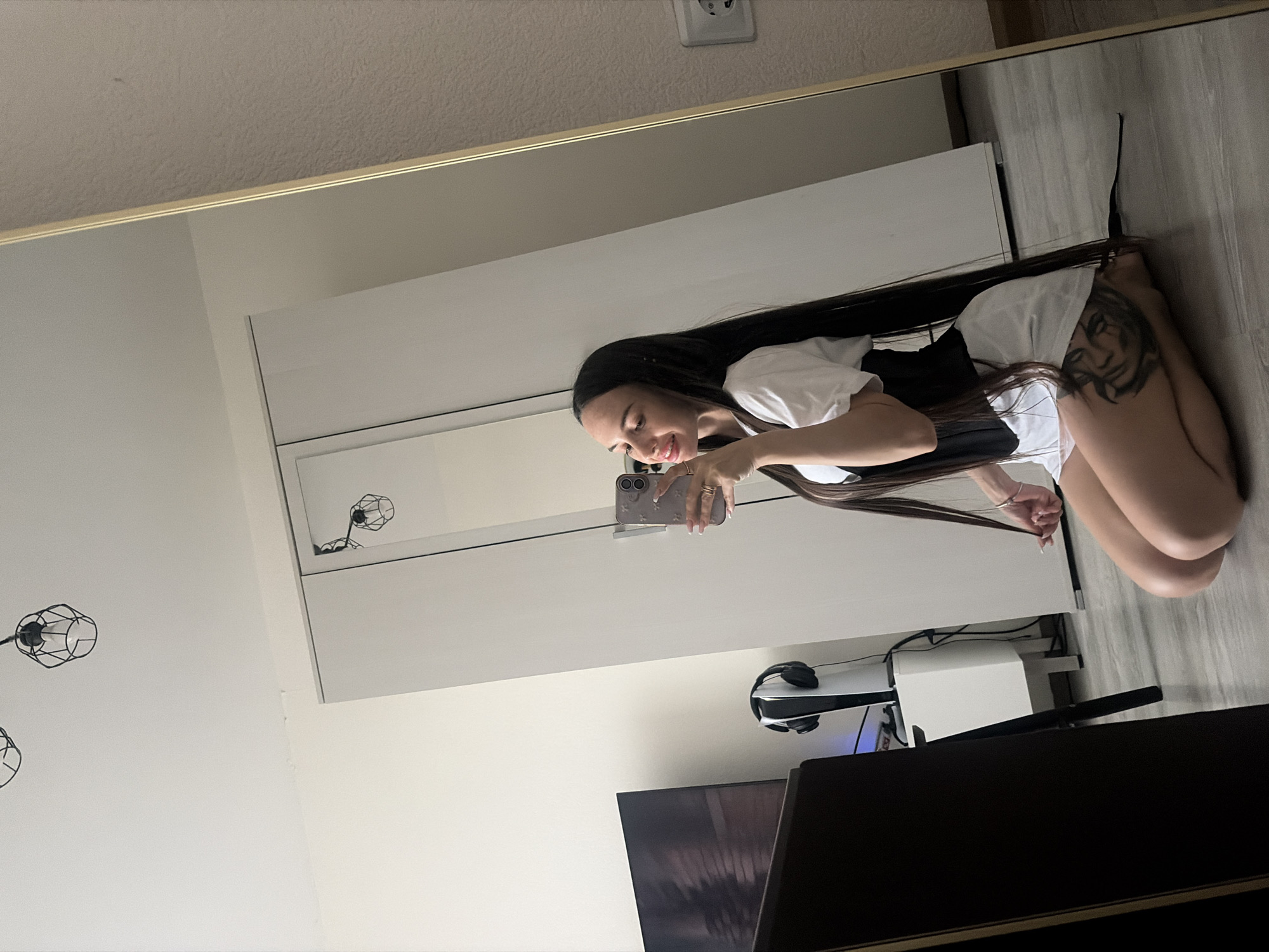 A mirror reflects a woman crouching in what appears to be a bedroom. She has long, dark hair and is wearing a white top and black shorts. A tattoo is visible on her right leg. She is taking a selfie with a smartphone. Reflected in the mirror is a white wardrobe or closet, a wall with a light fixture, a television, and a gaming console. The scene does not appear to be set in Saint Petersburg, Russia.