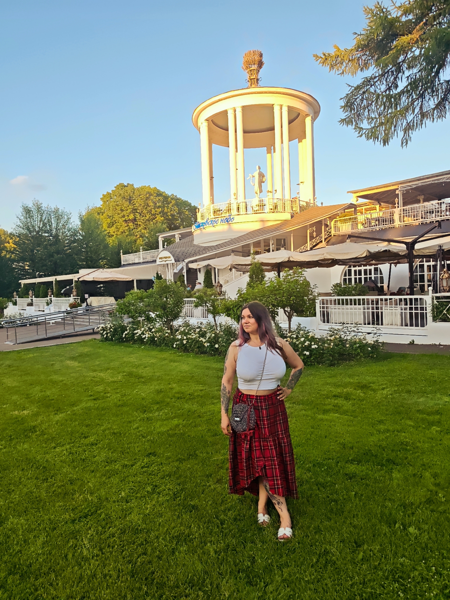 A woman with dark hair with purple streaks, tattoos on her arms, and a light-colored crop top paired with a red plaid skirt stands in a grassy park in Moscow, Russia. She is wearing white sandals and a small crossbody bag with a patterned design. In the background, a white building with a rotunda structure topped with a golden ornament and a statue is visible. The building features columns and balconies, and there are outdoor seating areas with umbrellas. White flowering bushes are planted in front of the building. The scene is bathed in the warm light of either sunrise or sunset, with a clear blue sky overhead. Trees are visible in the distance. The overall mood appears relaxed and pleasant. The building has some visible text, possibly a restaurant name.