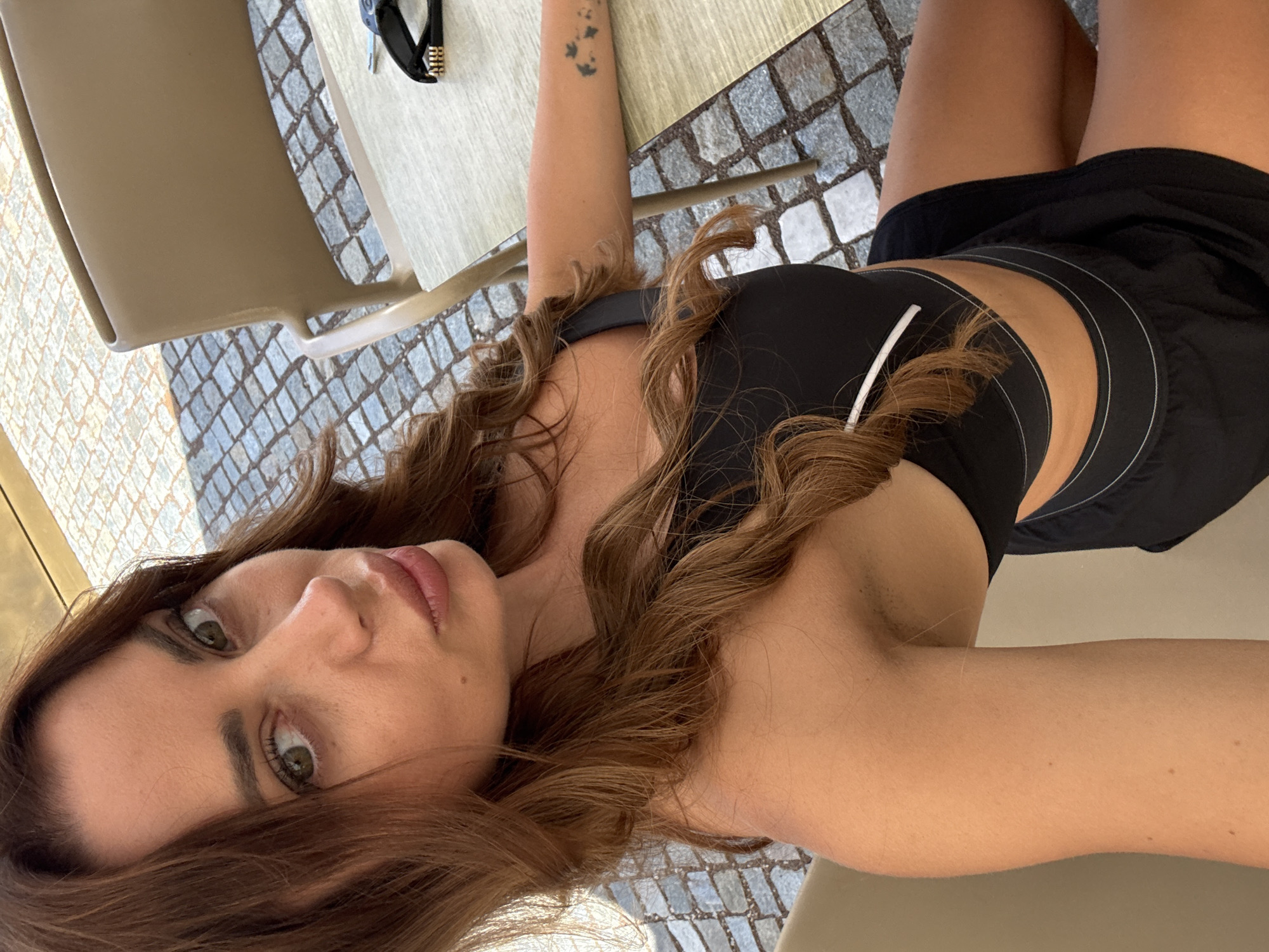 A woman with medium-length, wavy brown hair and green eyes is seated at an outdoor table with a light-colored surface. She is wearing a black sports bra and black shorts. Her left arm is resting on the table, and she has a small tattoo on her forearm. In the background, cobblestone pavement is visible, and a beige chair is positioned behind her. A pair of sunglasses and keys are on the table. The scene appears to be an outdoor seating area in a European city, potentially Calfosch - Kolfuschg - Colfosco, Italy, given the cobblestone and outdoor cafe setting. The lighting suggests it is daytime.