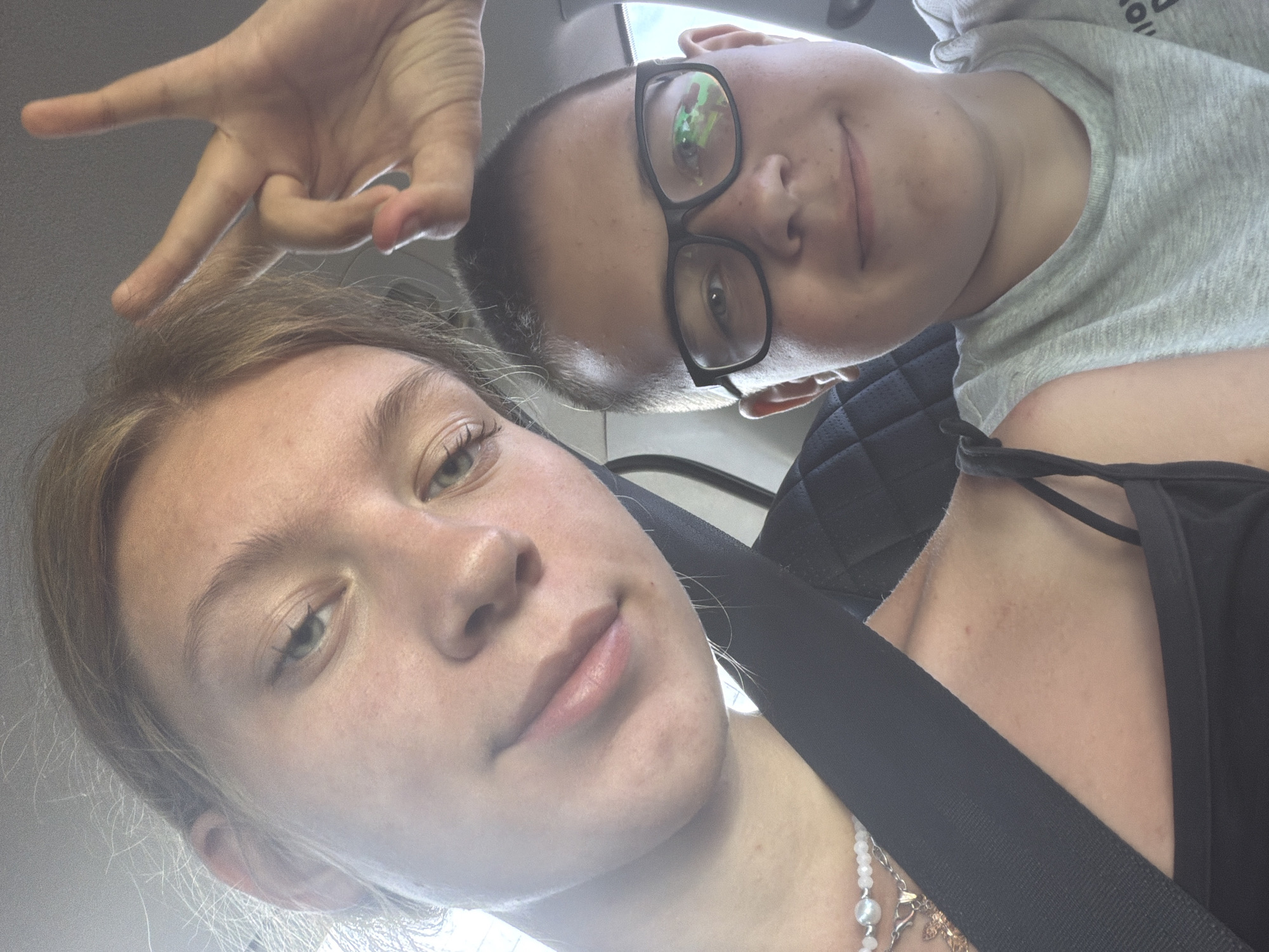 Two individuals are visible in a close-up selfie taken inside a vehicle. On the left, a young person with blonde hair is making a peace sign with their right hand. They are wearing a black tank top and a necklace with white beads and a pendant. Their left shoulder and chest area, including a seatbelt, are also visible. On the right, a young person wearing glasses and a grey t-shirt is looking towards the camera. The background shows the interior of a car, with a textured seat and a window. The image appears to be taken during daylight, possibly in Novoe Feizullovo, Russia, given the provided location context.