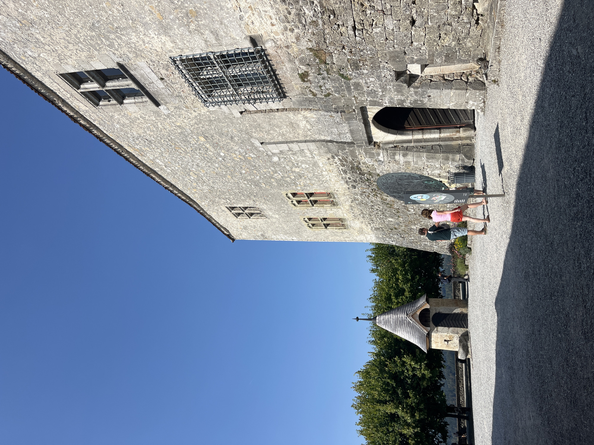 In the city of Epagny, Switzerland, on a clear sunny day, a stone castle stands tall against a bright blue sky. In the foreground, a gravel courtyard is partially shaded by the castle's imposing structure. To the left, a small, charming stone building with a conical wooden roof and a pointed spire sits nestled amongst lush green trees. A few people are visible in the courtyard: two individuals, a man and a woman, stand near the small building, seemingly engaged in conversation. Further to the right, two more individuals, a man and a woman wearing a pink shirt and red shorts, are walking towards the castle entrance. The castle's textured stone facade is adorned with several windows, some featuring intricate metal grates, hinting at its historical significance. The main entrance to the castle is an arched doorway, partially open, revealing a glimpse of what lies within.