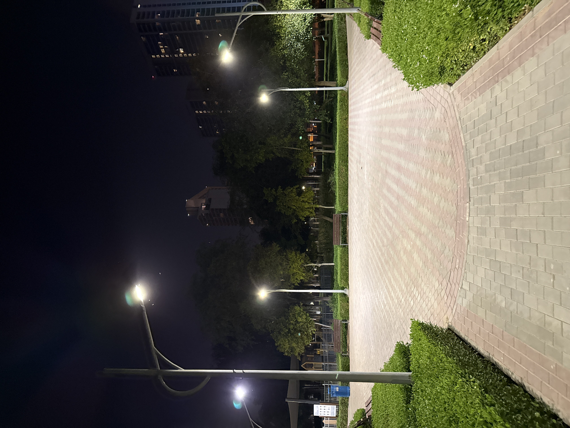 A night time, eye-level, wide shot of an empty park in Dubai, United Arab Emirates. The park is illuminated by streetlights, casting a warm glow on the brick pathway. Trees and manicured bushes line the path, creating a serene atmosphere. In the background, high-rise buildings are visible against the dark sky, suggesting an urban setting. Several park benches are placed along the pathway, inviting visitors to rest and enjoy the peaceful ambiance.