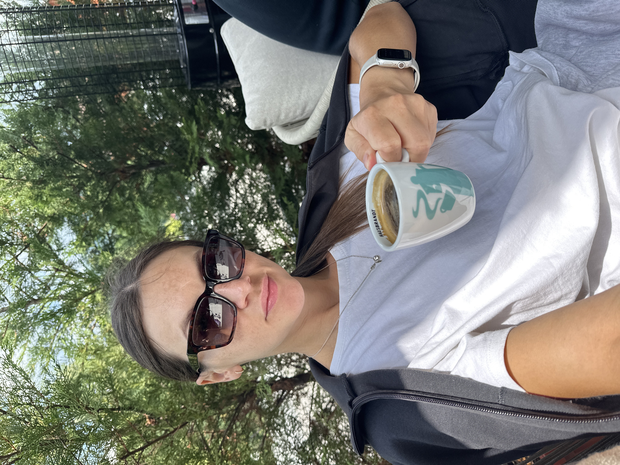 A woman sits outside, holding a mug of coffee. She is wearing sunglasses, a white t-shirt, and a black jacket. A smartwatch is visible on her wrist. The background includes trees and a patio heater, suggesting an outdoor seating area. The scene is set in Kolašin, Montenegro.