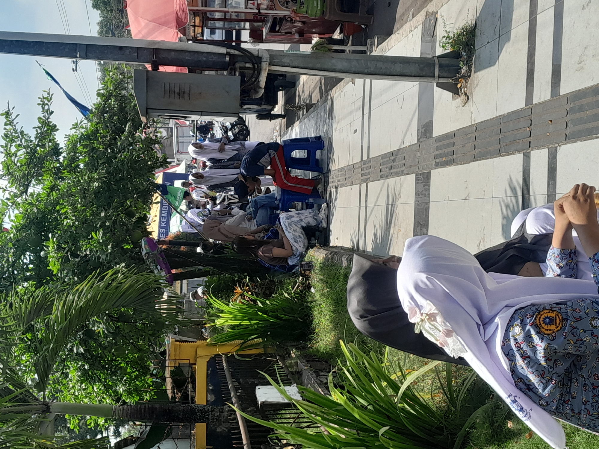 In Genteng Kulon, Indonesia, on a sunny day, a group of people are gathered along a paved sidewalk. In the foreground, two young women wearing hijabs are seated, one in a white hijab with decorative accents and a patterned uniform, and another in a black hijab. Behind them, several other individuals, many also in hijabs, are seated on blue plastic stools or on the ground. One person, wearing red pants and a dark t-shirt, is seated on a blue stool and appears to be looking down at their hands. In the background, lush green trees with palm fronds provide shade. A yellow fence and a black metal railing are visible along the edge of a grassy area. A gray electrical box is mounted on a pole near the sidewalk, and a red awning of a food stall is visible further in the background. The scene suggests a public gathering or waiting area, possibly near a marketplace or event.