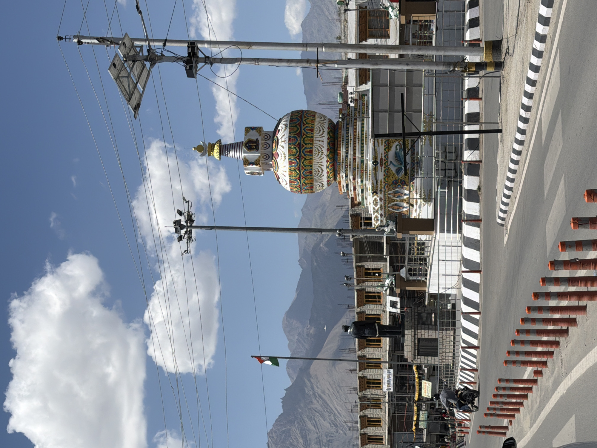 The image depicts a busy street scene in Leh, India, with a prominent stupa as the central focus. The stupa, a white dome-shaped structure adorned with colorful Buddhist motifs, stands tall against a backdrop of majestic, rugged mountains. In front of the stupa, a statue of a man in a uniform is visible, with an Indian flag flying beside it.
To the left of the stupa, buildings with traditional architecture, featuring wooden window frames and layered roofs, line the street. A billboard with text and a partial image is also visible on one of the buildings.
A road stretches across the foreground, with traffic barriers and orange and white striped bollards delineating the lanes. A motorcycle with a rider is seen on the road, moving away from the viewer. Several power poles with overhead wires and solar panels are scattered throughout the scene, indicating a mix of traditional and modern infrastructure.
The sky above is a clear blue, dotted with fluffy white clouds, and an airplane is visible flying in the distance, adding a sense of depth to the aerial perspective. The overall scene captures the vibrant cultural and urban landscape of Leh, showcasing its unique blend of tradition and modernity amidst its stunning natural surroundings.