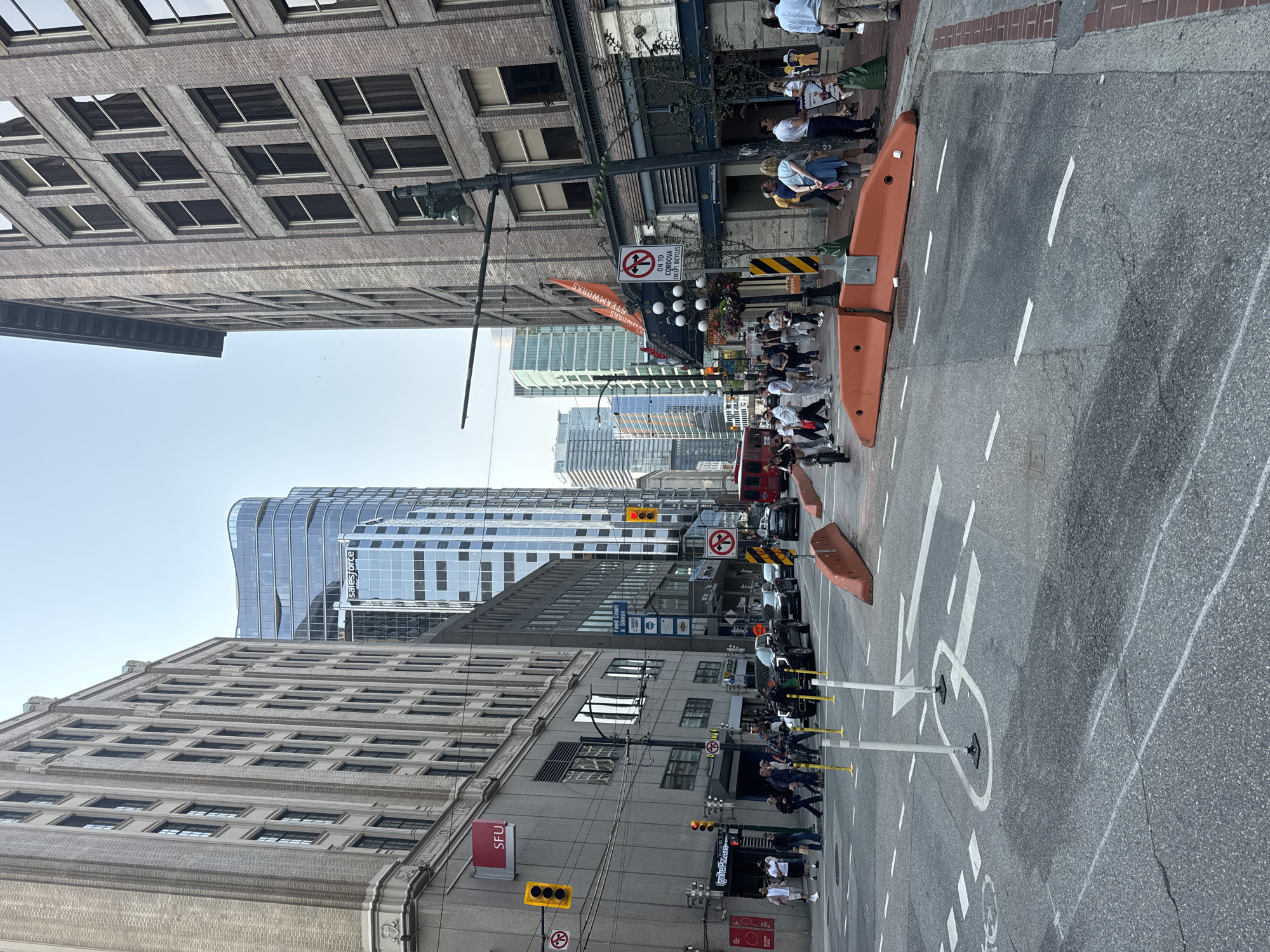 The image depicts a street scene in downtown Vancouver, Canada, likely near the intersection of Granville Street and West Cordova Street. Several multi-story brick and glass buildings line the street, characteristic of the Gastown or financial district areas. In the foreground, traffic cones and orange barriers are present on the asphalt road, indicating ongoing construction or traffic management. Pedestrians are walking on the sidewalks and crossing the street, some are wearing casual clothing. A red trolley bus is visible in the distance, and various vehicles are on the road. Signs are posted on buildings and poles, including business names and traffic instructions. The sky is clear, suggesting a daytime scene. The overall impression is a busy urban environment.