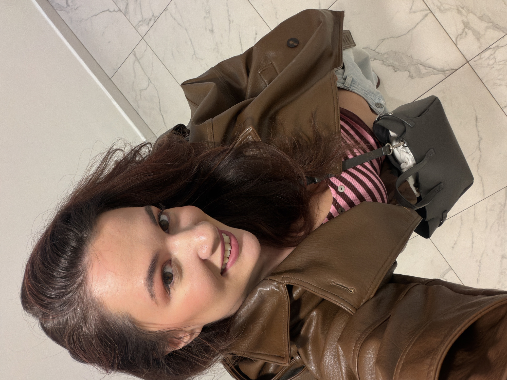 A woman with brown hair is smiling at the camera. She is wearing a striped cardigan and a brown leather jacket. A small gray purse is sitting next to her. The background appears to be a white tiled floor and a white wall. The location is not identifiable as Belgrade, Serbia based on the given information.