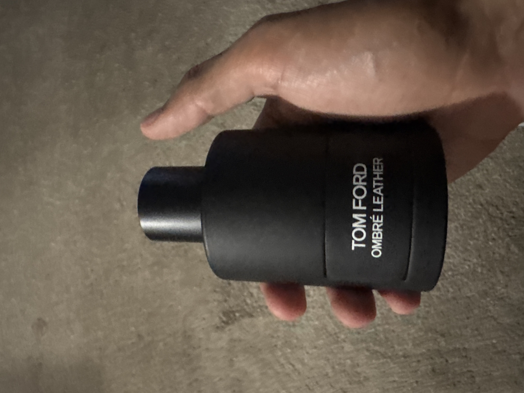 A human hand holds a black bottle of Tom Ford Ombré Leather fragrance. The background is a textured, neutral-toned surface. The location is Glendale Heights, United States of America.