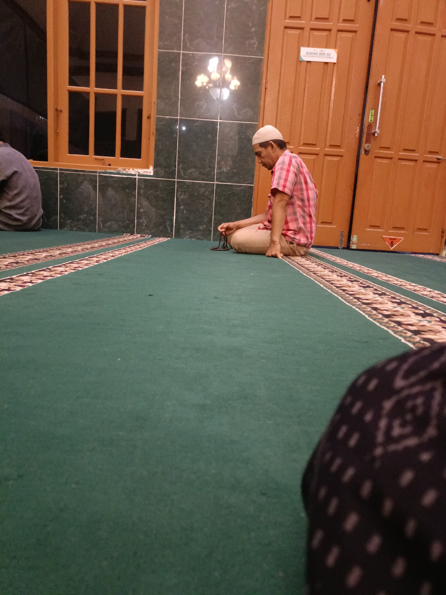 The image captures an indoor scene, likely within a mosque in Kediri City, Indonesia. A man, wearing a white skullcap and a red and white checkered shirt, is seated on a green carpeted floor. He is holding a string of prayer beads in his hands and appears to be engaged in prayer or contemplation. The floor is adorned with patterned carpet runners. In the background, there are wooden doors and a window with multiple panes. To the left, a portion of another person's back, wearing a dark grey shirt, is visible. The lighting suggests it is either dusk or the interior lights are on. The overall atmosphere is one of quiet reverence.