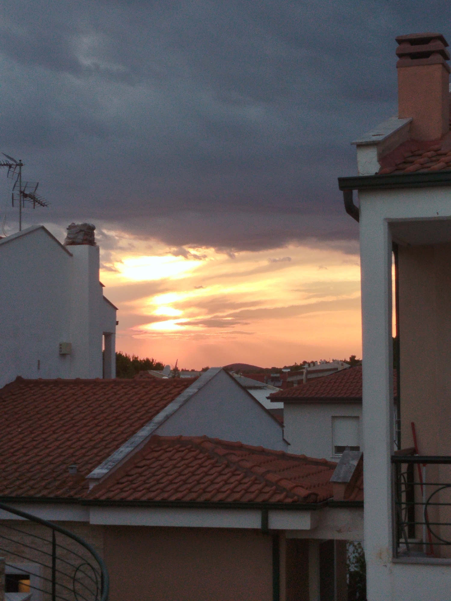 A vertical, medium shot captures a sunset over a coastal town in Sithonia Municipal Unit, Greece, as viewed from an elevated position. The sky is a dramatic mix of dark gray clouds in the upper portion and vibrant orange and yellow hues of the setting sun closer to the horizon. Several buildings with red tiled roofs are visible, some appearing to be multi-story residences. An outdoor antenna stands prominently on the roof of a white building on the left side of the frame. A brick chimney rises from a rooftop towards the upper right corner. The foreground is dominated by the steeply angled red-tiled roofs of multiple structures, with a portion of a white building and a balcony railing on the far right. In the distance, the silhouette of rolling hills can be seen against the bright sky. The scene appears to be devoid of people or any immediate activity.
