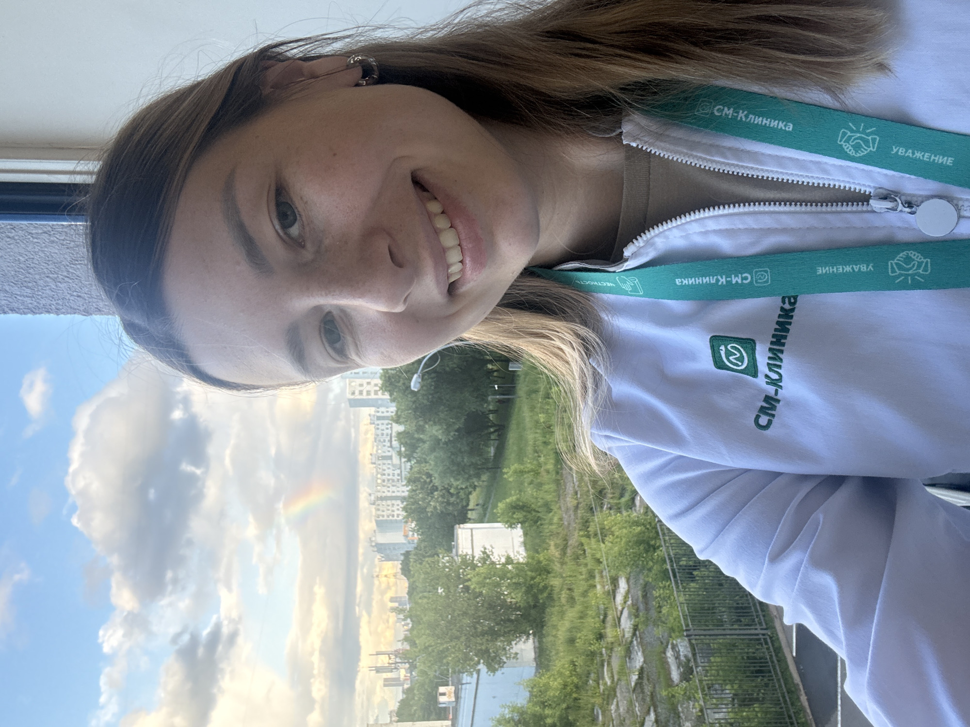 A young woman with long brown hair is taking a selfie outdoors. She is wearing a white zip-up jacket with green lettering that reads "СМ-КЛИНИКА" and has a green lanyard around her neck with the same text and images of shaking hands. The background shows a cityscape with buildings and trees under a cloudy sky with a rainbow visible. The image appears to be taken during the day. The woman has a light complexion, freckles, and is smiling. She is wearing hoop earrings. The woman is positioned in the foreground, with the cityscape taking up the midground and background. The scene suggests an urban environment in Moscow, Russia, possibly a residential or commercial area given the multi-story buildings and green spaces.