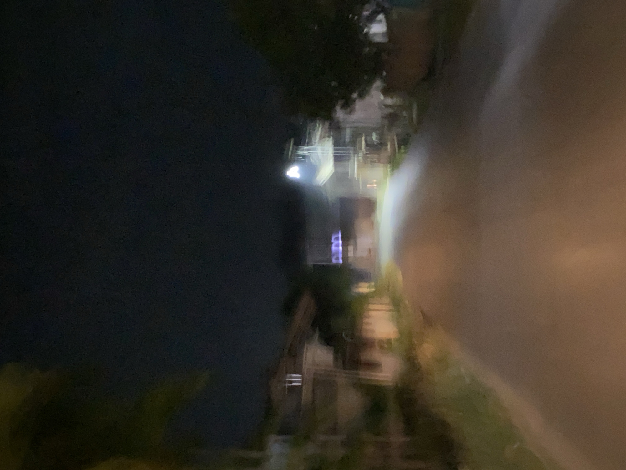 The image depicts a blurry, nighttime street scene in Lhokseumawe, Indonesia. A paved road stretches into the distance, flanked by vegetation and structures on either side. On the left, there are houses with indistinct features, one of which appears to have vertical white elements on its roof. On the right, a building with multiple levels is visible, illuminated by a bright light at its top. The sky is dark, indicating the absence of daylight. The overall impression is one of a residential or semi-commercial area at night, with limited visibility due to the blurriness and low light conditions.
