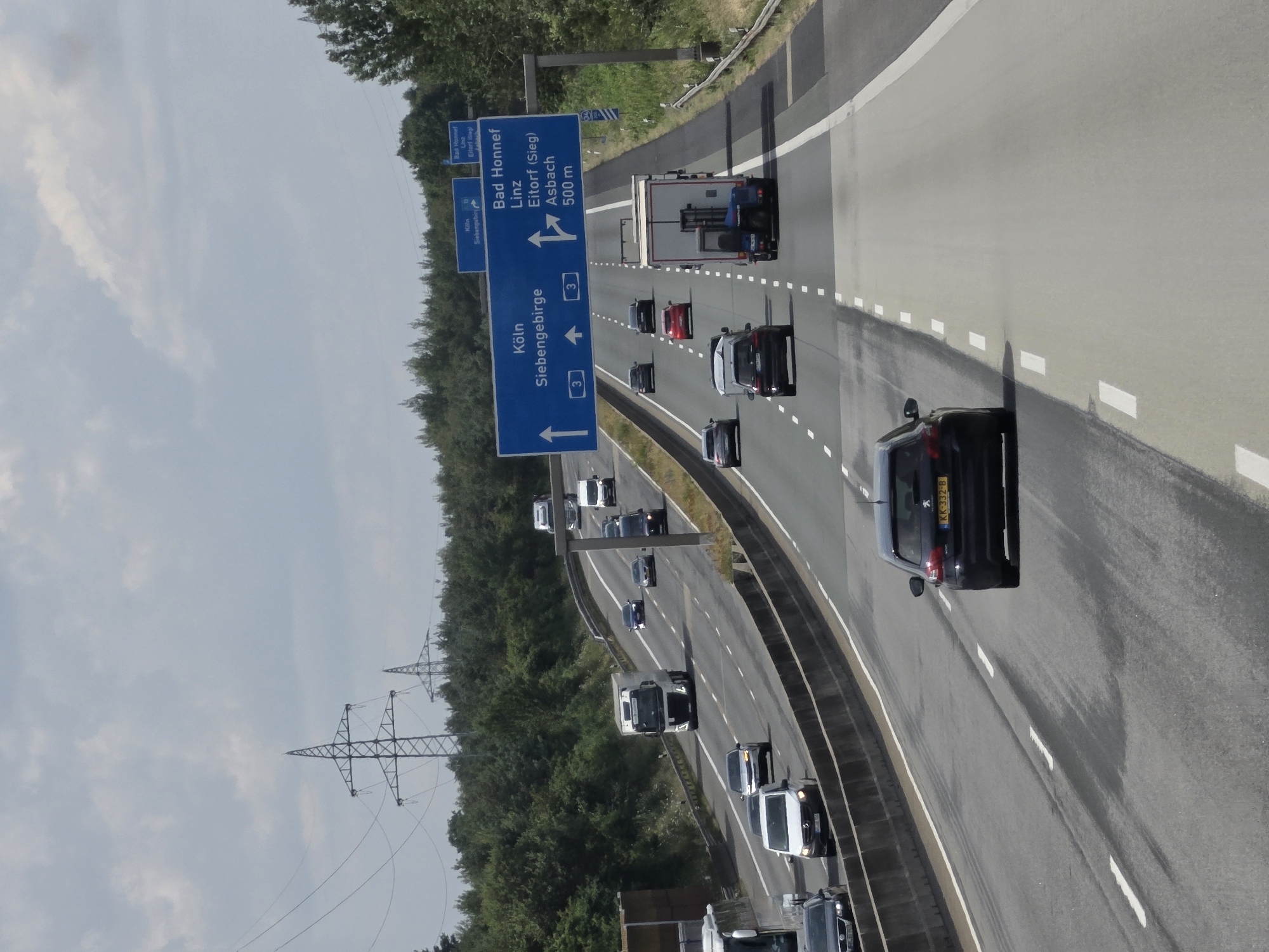 This is a full, eye-level shot of a multi-lane highway with several vehicles moving in both directions. The highway is flanked by trees on the left and right, with a concrete barrier separating the lanes. Power lines and transmission towers are visible in the background. The sky is overcast with some clouds.
Specifically, this image captures a section of the A3 Autobahn near Windhagen, Germany. A large blue overhead sign indicates directions to Köln (Cologne) and Siebengebirge straight ahead, and to Bad Honnef, Linz, Eitorf (Sieg), and Asbach 500m to the right. Several cars and a large white truck are visible on the highway. The license plate of the black Peugeot in the foreground is visible and reads "KK 332-B". The road surface shows signs of wear and some darker patches, possibly from recent rain.