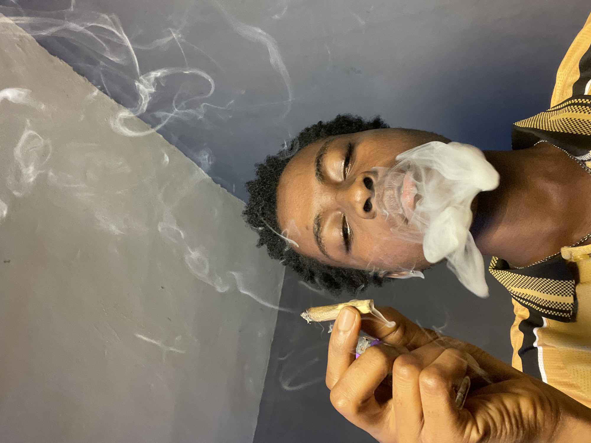A young Black man with short curly hair and closed eyes is exhaling smoke from his mouth, holding a lit marijuana cigarette in his right hand. He is wearing a yellow and black shirt with a checkered pattern. The background consists of a gray and off-white wall. The location is stated to be Nkpor, Nigeria.