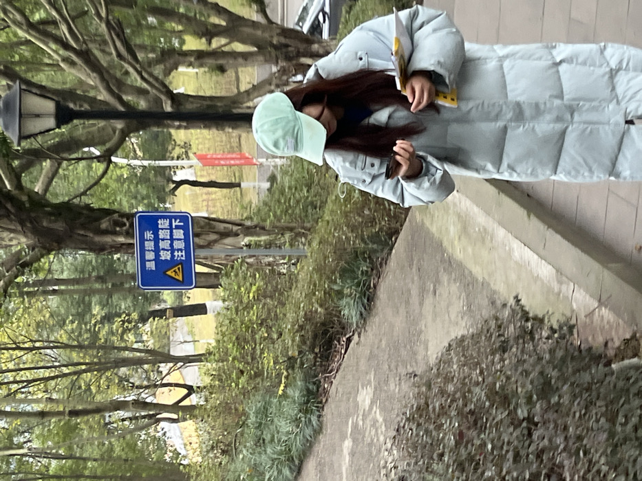 A person in a long, light blue puffer coat and a light green cap is walking on a sidewalk. The person is holding papers or a notebook. A sign warning of a steep slope is visible in the background. Trees with bare branches line the path. A lamppost stands near the sign. The scene is set outdoors, likely in a park or campus setting. The photo is taken during daylight.