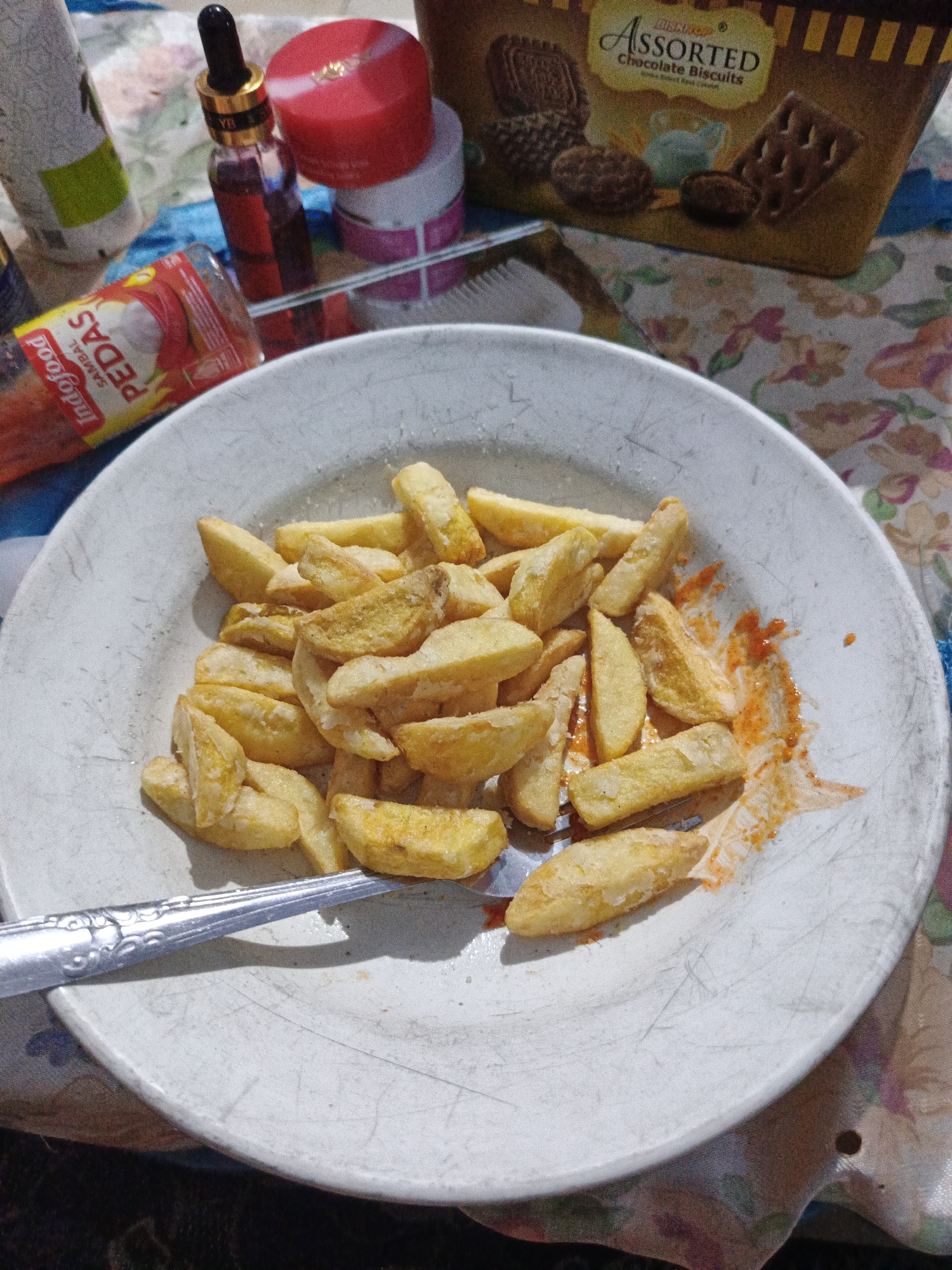 In a close-up shot, a white plate filled with golden-brown french fries is presented. The fries appear seasoned and are arranged in a pile, with some showing red sauce splattered on them. A silver fork rests on the plate, partially submerged in the fries. In the background, to the left, a red tube of "Indofood Pedas Sambal" is visible, along with a small dark bottle with a dropper and a red-lidded jar. To the right, a cardboard box with the text "Assorted Chocolate Biscuits" is partially in frame. The scene is set against a colorful floral-patterned tablecloth. The lighting suggests an indoor environment, likely a domestic setting within Sumbersari, Indonesia.