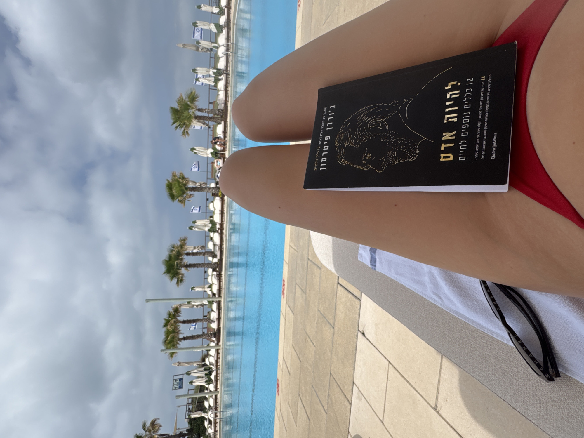 The image depicts a view of a person lounging by a pool, likely at a resort or hotel, given the presence of numerous lounge chairs, palm trees, and Israeli flags. A book titled "12 More Rules for Life: Beyond Order" by Jordan Peterson is positioned in the foreground, resting on the person's legs. A pair of sunglasses are visible on a towel near the legs. The sky is cloudy, suggesting overcast weather.
While the location is not explicitly mentioned, the presence of Israeli flags suggests it could be in Israel, potentially in the city of Herzliya.