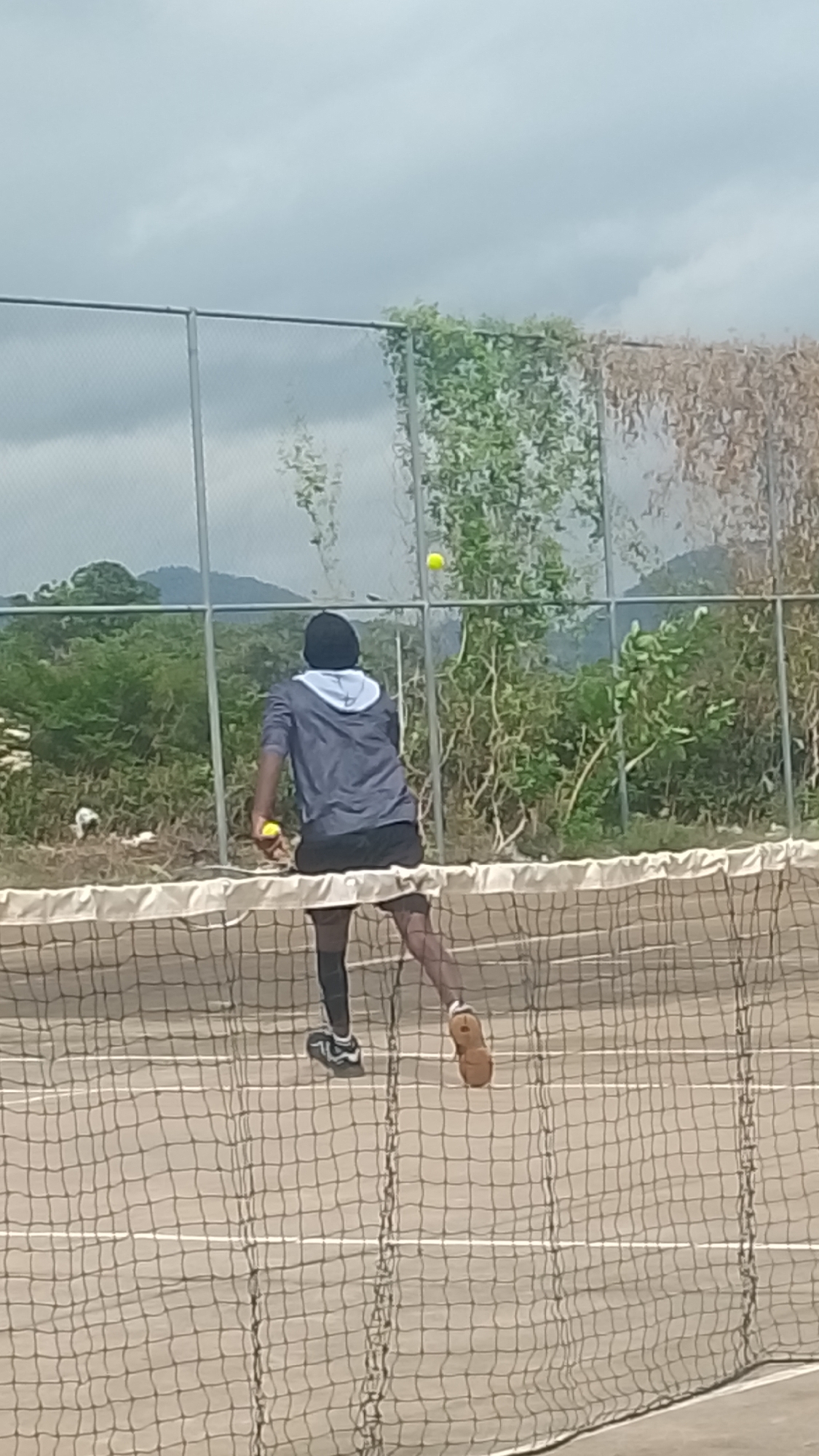 The image depicts a tennis court in Lokoja, Nigeria, viewed from behind the net. A single individual, seen from the back, stands on the far side of the court, facing away from the camera. The person wears a dark long-sleeved top with a light-colored hood, dark shorts, and a dark compression sleeve on the left leg, paired with athletic shoes—one dark with white accents on the left foot and a brown shoe on the right. A yellow tennis ball is held in the person's left hand. Another yellow tennis ball is suspended in mid-air above the court on the far side of the chain-link fence. The court surface is a light brown or grey, marked with white lines partially obscured by the foreground tennis net. Behind the court and fence, dense green and some dry brown vegetation are visible, with distant mountains or hills under an overcast sky.