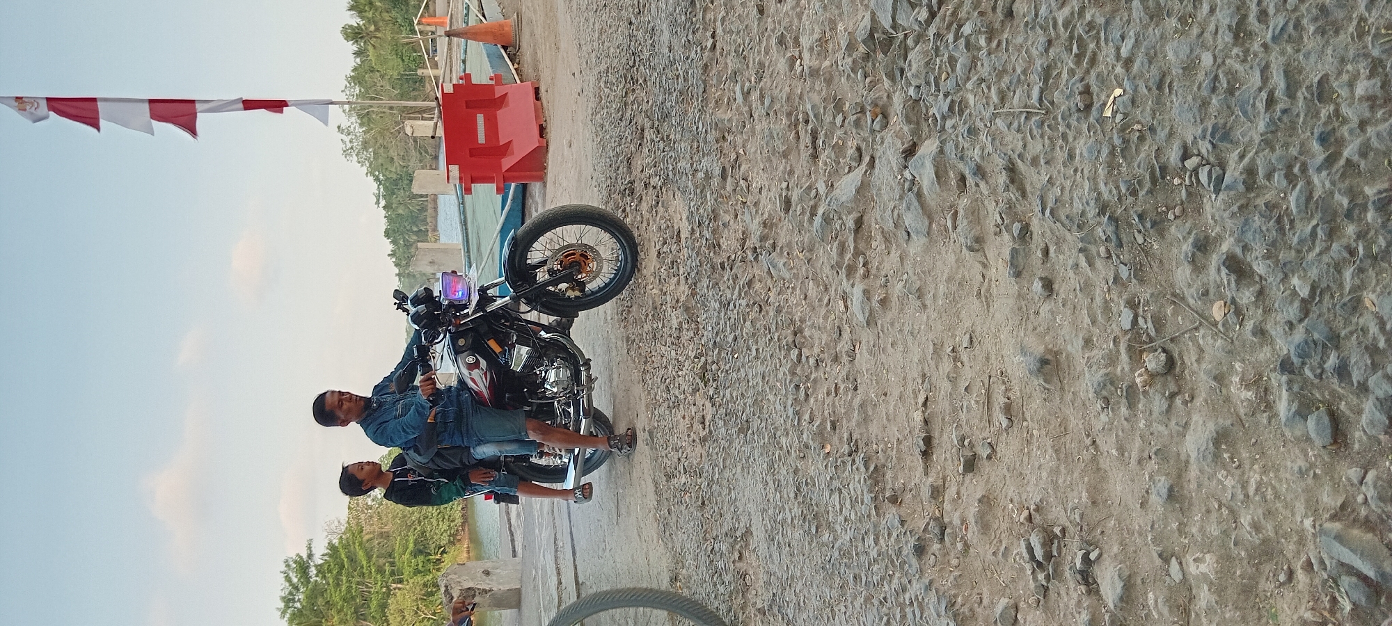 Two individuals, an adult male and a young male, are seated on a black motorcycle. The adult male is driving, while the young male sits behind him. Both are wearing casual clothing. The motorcycle has a red and black design on its side. They are positioned on a rough, gravelly surface that appears to be a road or pathway. In the background, a body of water is visible, lined with trees and vegetation. A red traffic barrier and an orange traffic cone are present on the right side of the scene. A partially visible Indonesian flag, with red and white stripes, is affixed to a pole on the far right. The sky above is clear with some wispy clouds. The location is identified as Kalipucang, Indonesia.