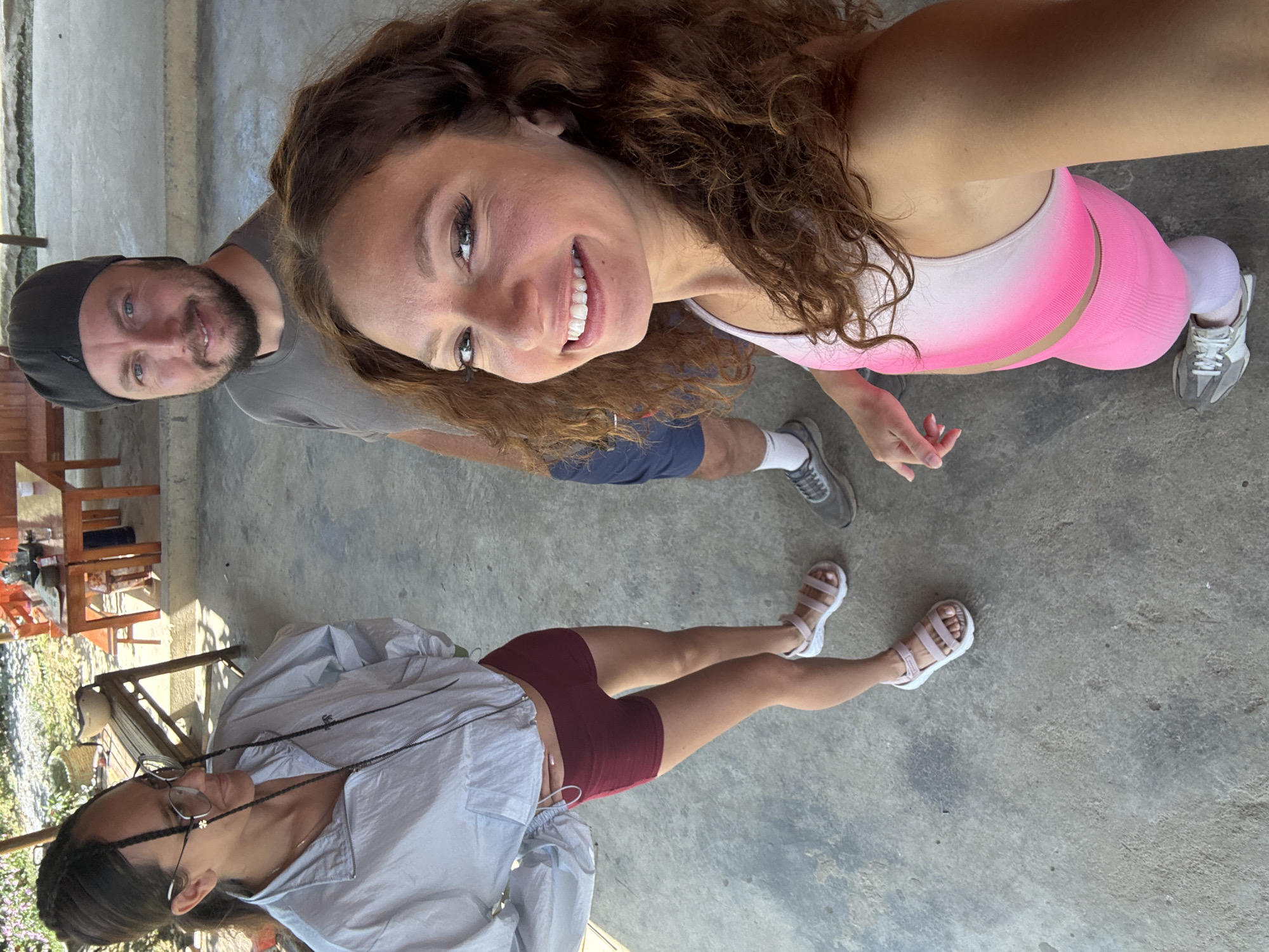The image is a selfie-style, aerial shot, presumably taken in Bắc Hà, Vietnam, featuring three individuals on a paved outdoor surface. The primary subject, a woman with curly brown hair, faces the camera with a smile. She wears a white-to-pink gradient sports bra and matching pink leggings, paired with gray athletic shoes and white socks.
Behind her and slightly to the left, a man with a beard and a dark cap looks into the camera. He is wearing a gray shirt and dark blue shorts, with gray sneakers on his feet.
Below them and angled towards the left edge of the frame, another woman lies on the ground. She is wearing a light gray button-down shirt, maroon shorts, and white sandals. She has dark hair and glasses and appears to be facing away from the camera.
The background suggests an outdoor setting. Hints of vegetation and what appear to be outdoor furniture (wooden tables and chairs) are visible. The ground surface appears to be concrete. The scene is brightly lit.