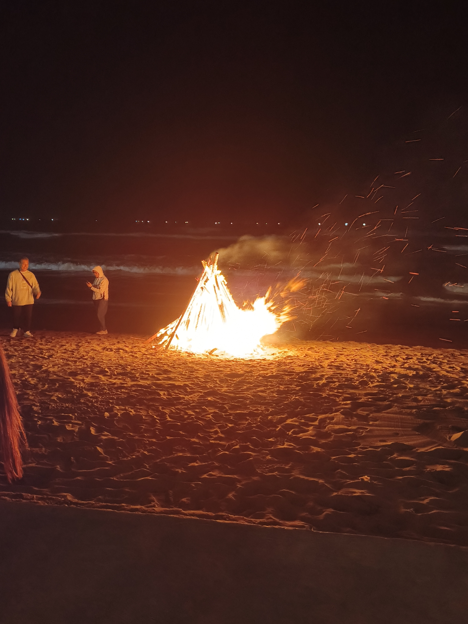 An outdoor nighttime scene captures a large bonfire on a sandy beach, with the ocean visible in the background. Two individuals are present on the beach. The person on the left, facing the bonfire, wears a light-colored hooded sweatshirt and dark pants, with white sneakers. The person on the right, also facing the bonfire, is dressed in a white hooded sweatshirt and dark pants, and is holding a mobile phone. The bonfire, constructed of stacked logs, is ablaze and casting a bright orange glow across the sand and the surrounding area. Sparks are visibly flying from the fire. The ocean in the background appears dark, with gentle waves. Distant lights, possibly from a coastline or boats, are faintly visible on the horizon. The beach sand is textured with small dunes and undulations. The image is captured from a low angle, emphasizing the height of the bonfire and the expanse of the beach. The overall atmosphere suggests a communal gathering or a moment of leisure by the sea.