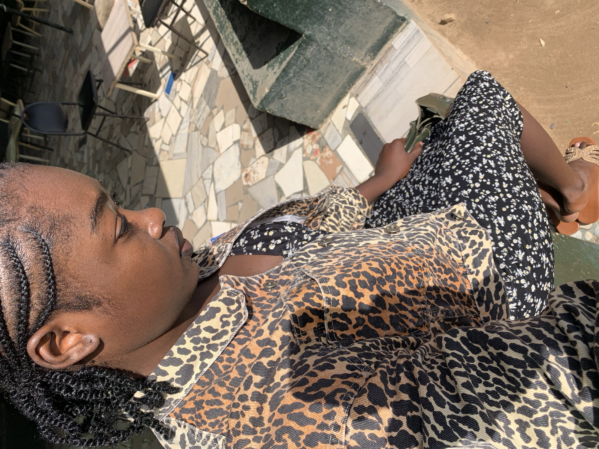 A person is captured in a sun-drenched outdoor setting, likely a patio or courtyard in Kabong, Nigeria. The image is taken from a slightly elevated perspective, looking down. The subject, a person with dark skin and braided hair, is seen from the side, with their eyes gently closed, suggesting a moment of relaxation or rest.
They are dressed in a distinctive leopard print jacket or shirt worn over a black dress or skirt adorned with a small white floral pattern. A dark green bag rests on their lap. On their feet, they wear light brown sandals embellished with gold-colored decorative elements.
The background features a tiled floor with a mosaic-like pattern of irregular light-colored and dark-colored pieces. Shadows cast by furniture, including chairs and possibly a table, are visible on the floor, indicating bright, direct sunlight. The overall impression is one of a calm, peaceful moment enjoyed outdoors on a sunny day. No text is visible in the image.