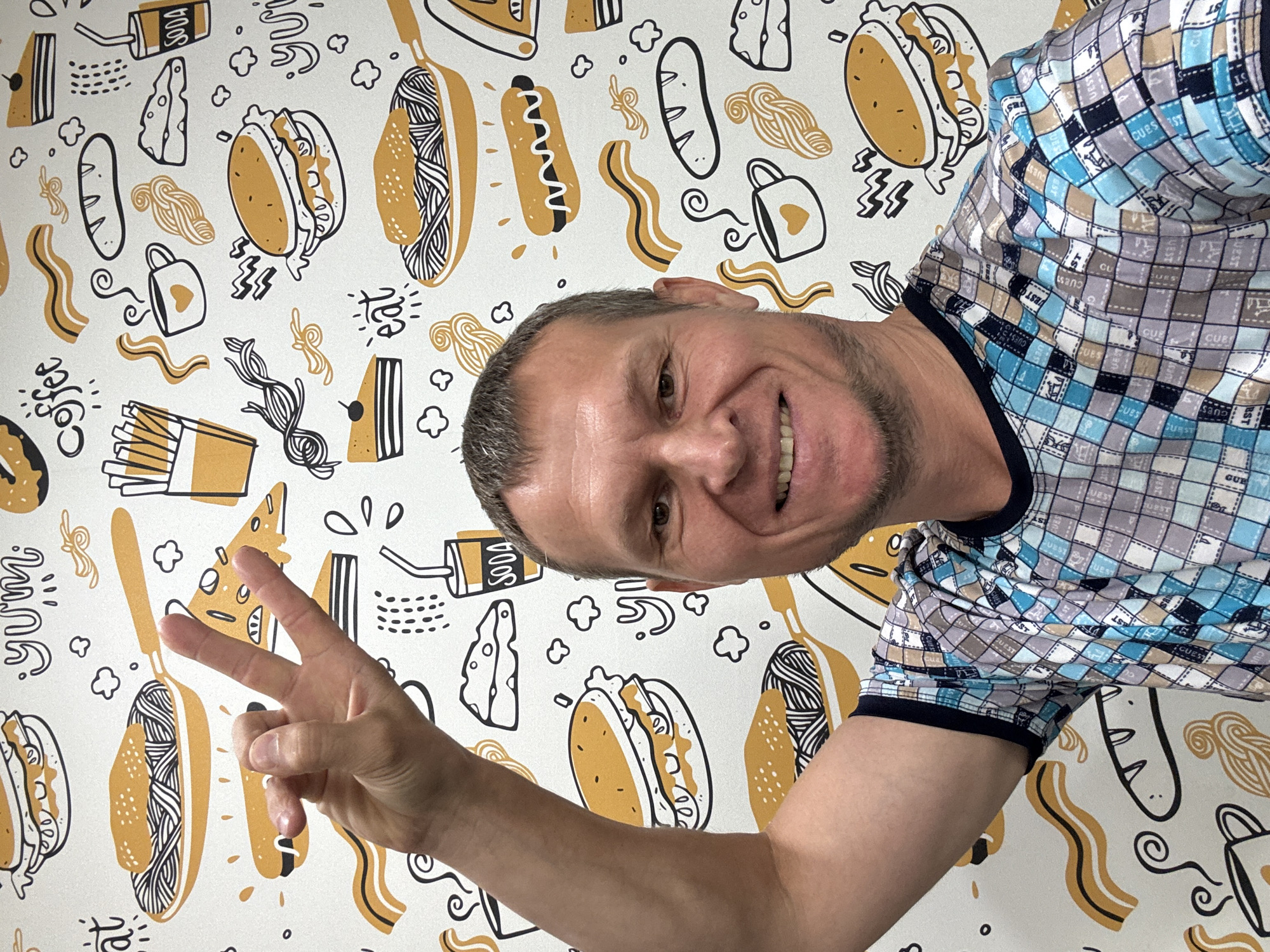A middle-aged Caucasian man is taking a selfie in front of a wall covered in a food-themed mural. The mural features illustrations of fast food items like burgers, fries, hot dogs, and coffee cups, as well as Italian food like pasta and pizza. The man is smiling and making a peace sign with his left hand. He is wearing a patterned t-shirt with blue, grey, and white squares. The background appears to be indoors, with the wall being the only visible environmental element. There is no information in the image that indicates the location in the city of Oskemen, Kazakhstan.