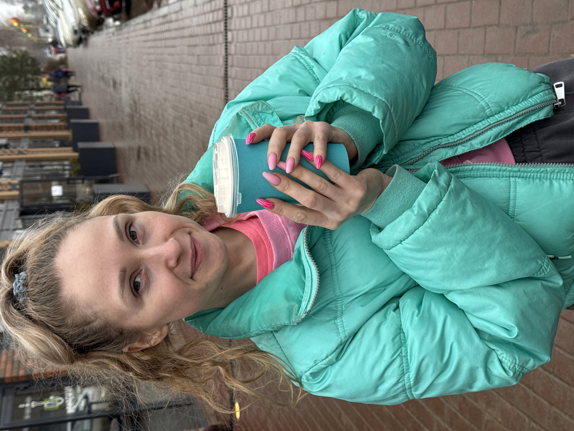A woman with blonde hair is holding a blue cup with a white lid, standing on a brick-paved sidewalk in Osa, Russia. She is wearing a light green puffer jacket and a pink sweater. Her nails are painted pink with a stripe design. In the background, there are rows of gray planters and buildings visible.