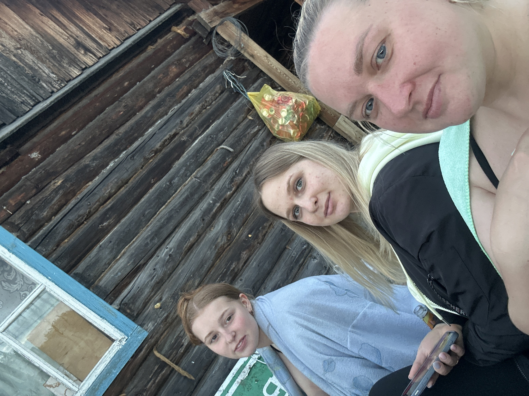 The image depicts three adult females posing for a photograph in Chaikovsky, Russia, positioned closely against the diagonal backdrop of a rustic, weathered wooden log house.
In the upper left portion of the frame, an old window with a distinct blue frame and white sash is visible. The window contains multiple glass panes, and through it, patterned wallpaper and a wooden board are discernible. A partial green sign is present on the left side, adjacent to the leftmost individual.
The upper right portion of the background features a translucent yellow plastic bag containing several small bottles, suspended from a wooden beam. Dark electrical wires are coiled and tangled near this bag and beam. The log wall itself consists of horizontally stacked, aged, dark wooden logs.
The leftmost female has reddish-brown hair styled in a bun and is wearing a light blue bathrobe with faint patterns. She looks directly at the camera. The central female has long blonde hair, wears a black jacket over a light-colored hooded garment, and holds a dark-colored smartphone in her left hand. A small, clear bottle with a light-colored cap is visible near her right hip. Her facial skin exhibits multiple small blemishes. The rightmost female, partially visible and cropped, has light blonde hair, blue eyes, and noticeable freckles on her fair skin. She is wearing a black strap garment, possibly a swimsuit or bra, and has a light green towel or piece of fabric draped over her chest and shoulder. All three individuals are looking forward, directly at the camera.