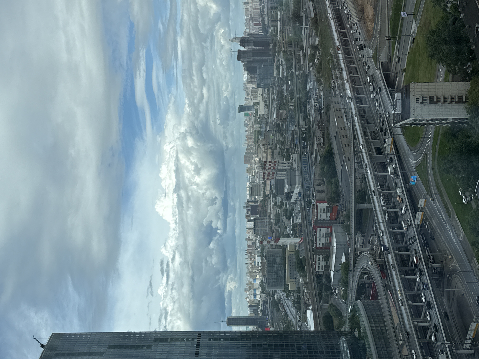 The image depicts a cityscape viewed from a high vantage point, with a significant portion of the frame occupied by the sky filled with thick, grey and white clouds interspersed with patches of blue. Below the clouds, a sprawling city extends into the distance, marked by numerous buildings of varying heights and architectural styles.
A complex network of roadways, including multi-lane highways and intricate interchanges, is visible on the right side of the image, with numerous vehicles traversing these routes. To the left of the roadways lies a dense urban landscape, composed of residential and commercial buildings. The foreground includes parts of the building from which the picture was taken.
While specific details about the people are indiscernible due to the distance, the scene suggests a bustling urban environment. The city in the image is Khimki, Russia, identifiable by its unique skyline and transportation infrastructure.