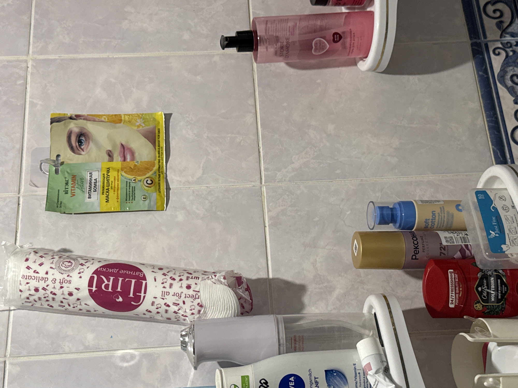 The image shows a collection of personal care products on a tiled wall in a bathroom. On the top left, a large pack of "Flirt" cotton pads is hanging from a hook. To its right, a single-use face mask packet labeled "Viteks Vitamin Active" is also hanging. Below these, on a white shelf, are various bottles and tubes of toiletries, including a white dispenser, a bottle of "NIVEA" body lotion, and a tube of hand cream. On a lower shelf, several more items are visible: a gold-capped spray bottle labeled "Рексона," a blue-capped spray bottle labeled "soft cotton," a red deodorant stick with "Old Spice" branding, and a small clear plastic container with what appear to be dental floss picks. To the right, on another shelf, is a pink bottle of "STORIE D'AMORE" body wash. The tiled wall and the arrangement of products suggest a domestic bathroom setting. The location is specified as Яромінскі сельскі Савет, Belarus.