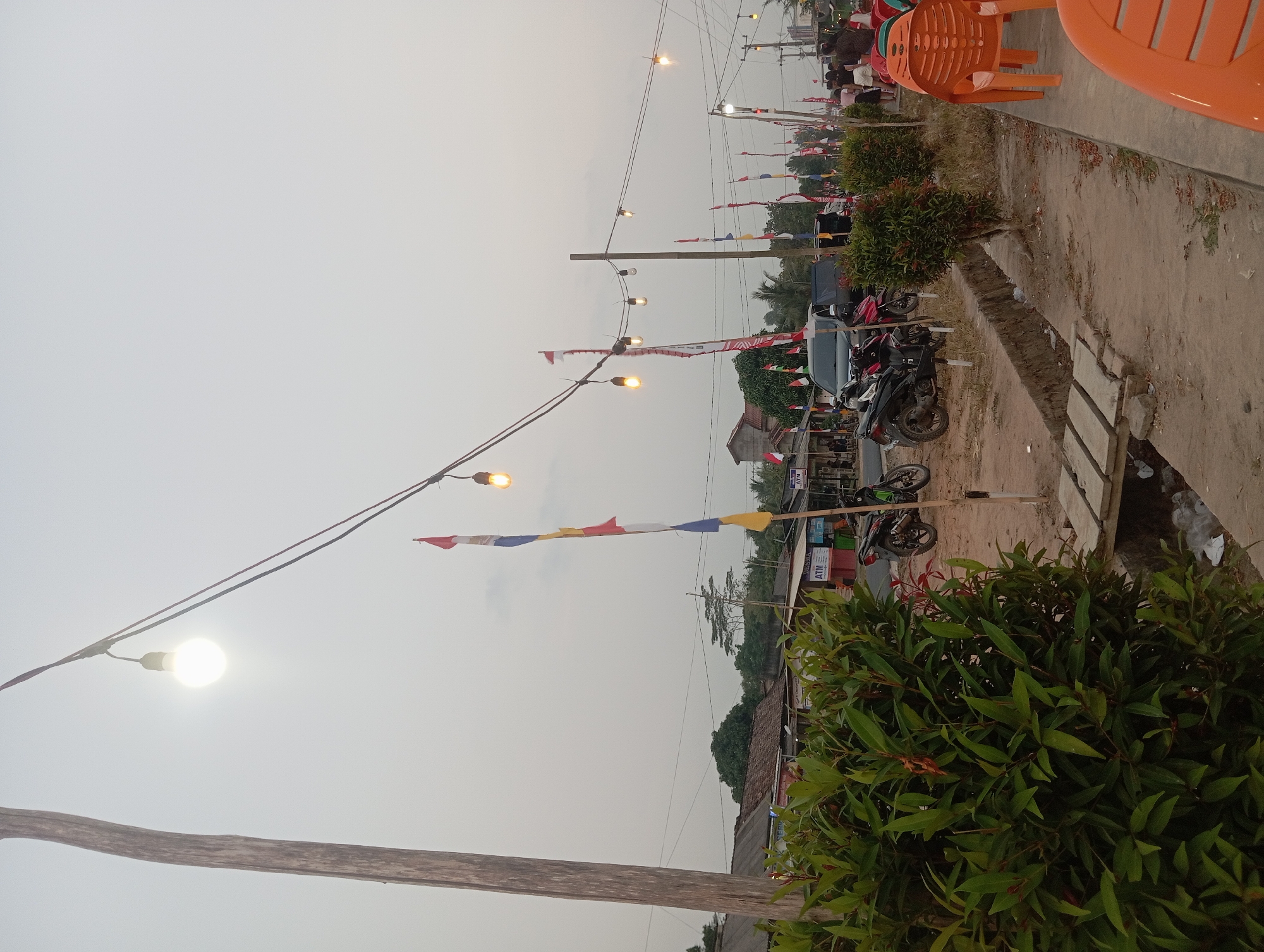 The image depicts an outdoor scene in Way Dente, Indonesia, likely during an evening event or market. In the foreground, a lush green bush is prominent on the left. To the right, several orange plastic chairs are arranged, suggesting a seating area for patrons.
The middle ground shows a dirt pathway with a small concrete channel running alongside it. Several motorcycles are parked on the dirt area, some appearing to be on a stand. A dark SUV is also visible parked in the background. Numerous Indonesian flags, a mix of red and white, are displayed on poles along the pathway, indicating a local celebration or national holiday.
Overhead, strings of bare light bulbs are suspended, some illuminated, casting a warm glow. The sky is overcast and pale. In the background, various buildings, likely shops or stalls, line the street. A few people are visible in the distance, milling about near the stalls and chairs. The overall atmosphere suggests a community gathering or a lively marketplace.
