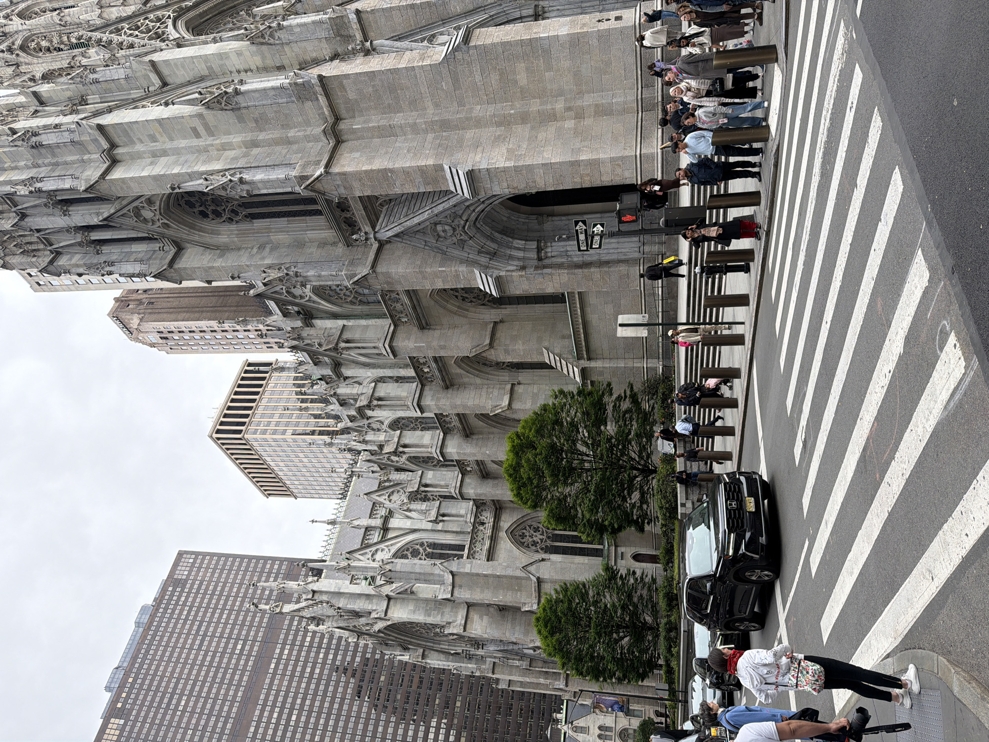 The image captures the iconic St. Patrick's Cathedral in New York City, United States of America. The Cathedral's facade is adorned with intricate architectural details, contrasting with the modern skyscrapers surrounding it. A crosswalk in front of the Cathedral is visible, with pedestrians waiting to cross. Cars are parked on the street, adding to the urban landscape. Green trees provide a touch of nature amid the city's concrete and steel. The scene conveys a sense of urban activity and historical grandeur coexisting in the heart of New York City.