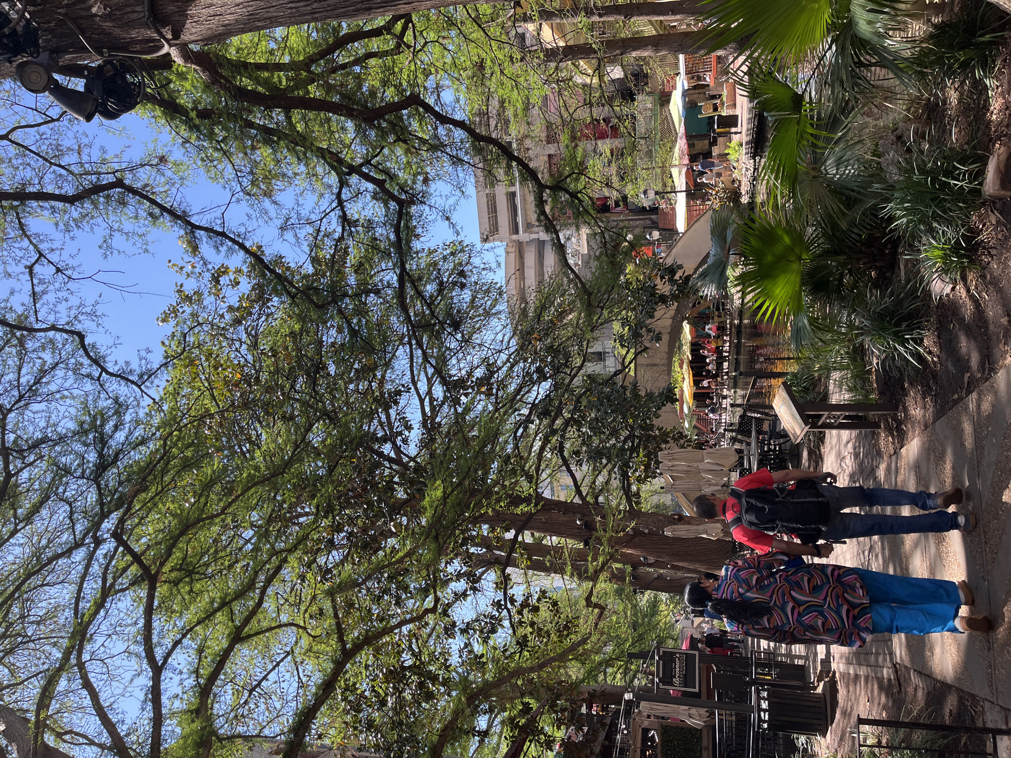 This image shows a section of the San Antonio River Walk in Texas, USA. In the foreground, two pedestrians walk along the paved walkway. One person wears a colorful patterned shirt and blue pants, while the other wears a red shirt and jeans, with a backpack. The walkway is bordered by metal railings and planters with vegetation. Beyond the walkway, there is a canal area, likely part of the San Antonio River, with outdoor dining areas visible. Tables with umbrellas and people seated are scattered along the waterfront. Buildings are visible in the background. Overhead, trees with green foliage provide shade. A lighting rig is secured in one of the trees.
