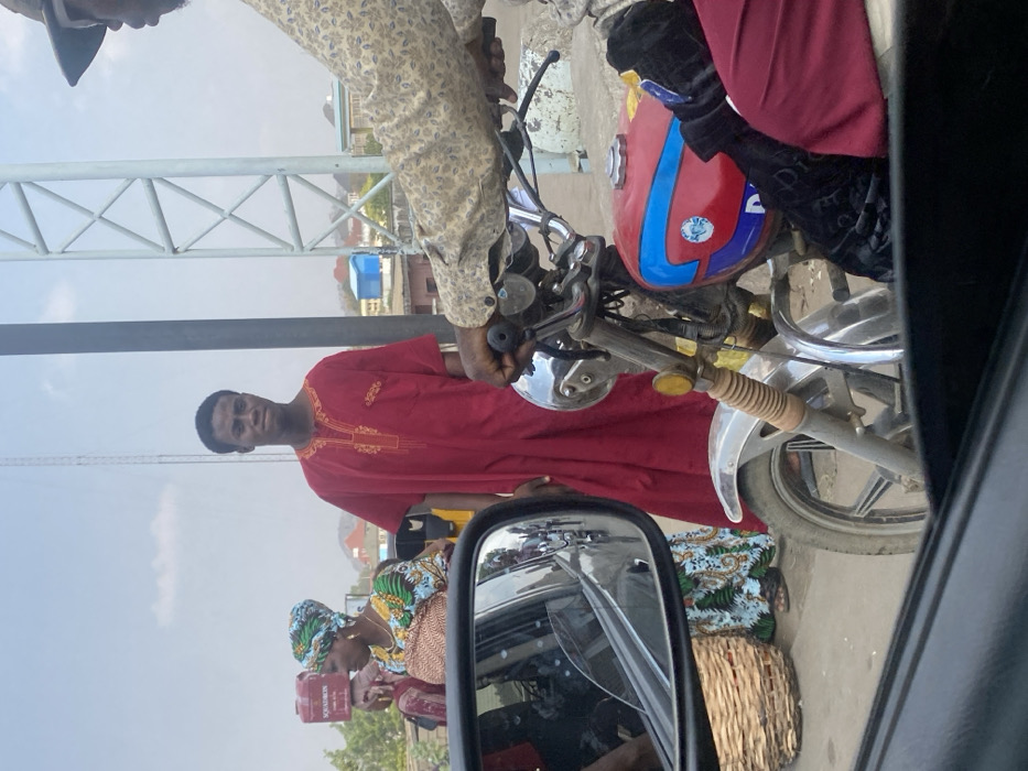 A man in a red tunic stands near a parked motorcycle with a red and blue fuel tank. Another person wears a patterned shirt while sitting on the motorcycle. A woman wearing a patterned headscarf and carrying a basket is visible near the side mirror of a vehicle where the photo is being taken. The background features buildings and a clear sky.