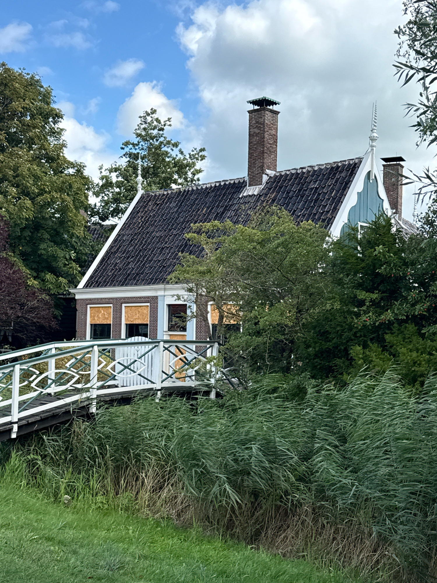 The image depicts a traditional Dutch house in Zaandam, Netherlands, characterized by its steep, dark-tiled roof and brick facade. Two prominent brick chimneys rise from the roofline, one with a decorative green cap. The house features distinctive white decorative gables on its upper facade. Some windows are covered with what appear to be rolled-up blinds or shutters, displaying an orange hue. A white and green lattice bridge with decorative elements spans a waterway or ditch in front of the house. The surrounding environment is lush and green, with tall reeds growing along the water's edge and mature trees flanking the property. The sky above is a mix of blue and white clouds. No people are visible in the scene.