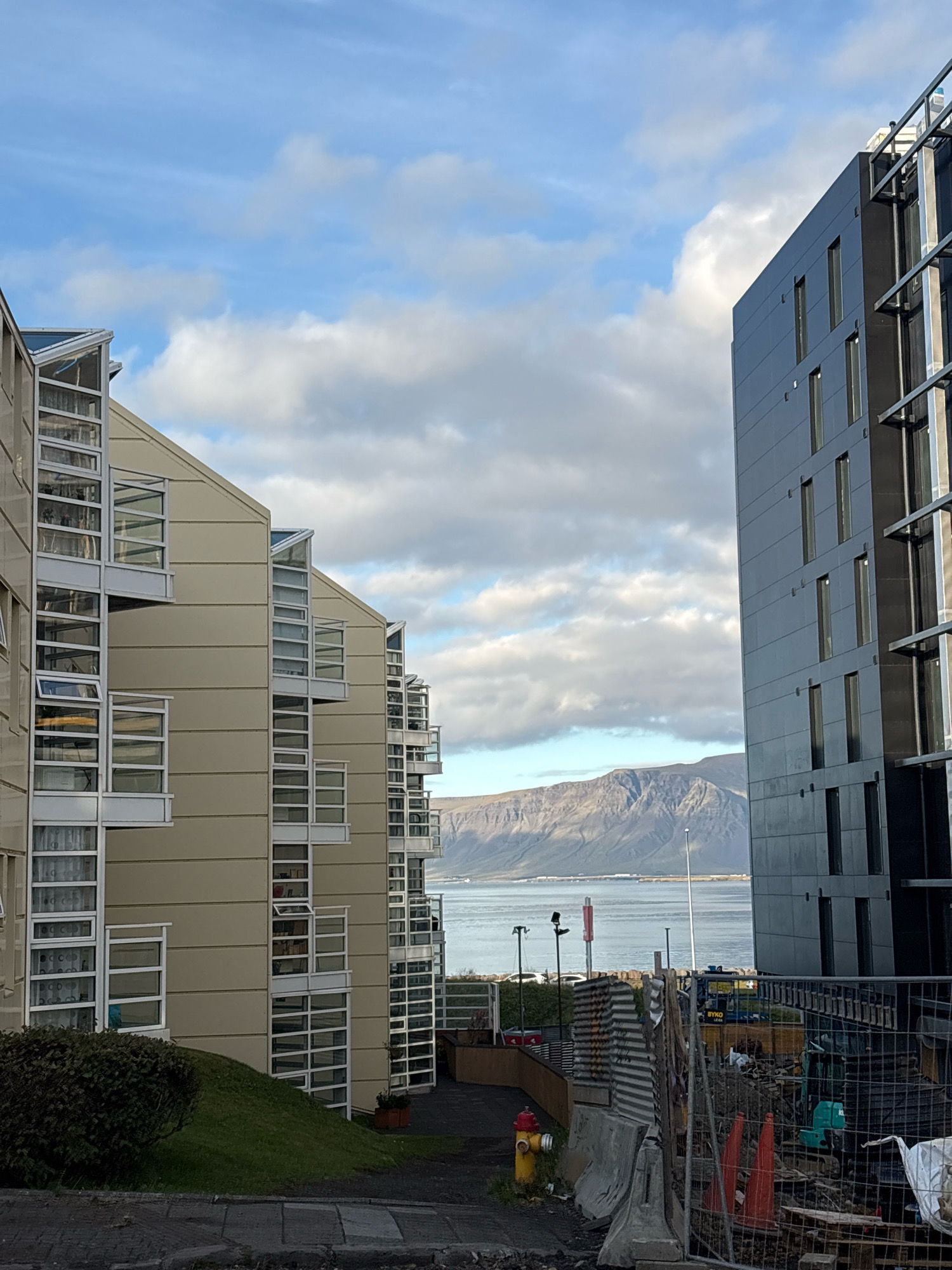 The image captures a view in Reykjavik, Iceland, featuring two modern buildings with distinct architectural styles flanking a body of water and a mountainous landscape in the background.
On the left, a beige-colored building with a repeating pattern of multi-story windows with white frames and some angled glass sections suggests residential or office units. These windows are staggered and project outwards, creating visual interest. Lush green grass and a small, dark green bush are visible in the foreground to the left, bordering a paved area.
On the right, a tall, dark, modern building with a facade of dark, rectangular panels dominates the frame. It has a series of vertically aligned, narrow windows. Adjacent to this building, and in the middle ground, is a construction site enclosed by a chain-link fence. Construction materials such as concrete barriers, orange traffic cones, and a small green excavator are present within the fenced area.
In the background, a calm body of water, likely a bay or harbor, stretches across the scene. Beyond the water, a large, rugged mountain range with distinct peaks and slopes forms the horizon. The sky is partly cloudy with patches of blue sky visible, indicating daytime. A lamppost and a red and white pole with signage are also visible near the waterfront. The overall impression is one of urban development meeting natural landscape, with a touch of ongoing construction.