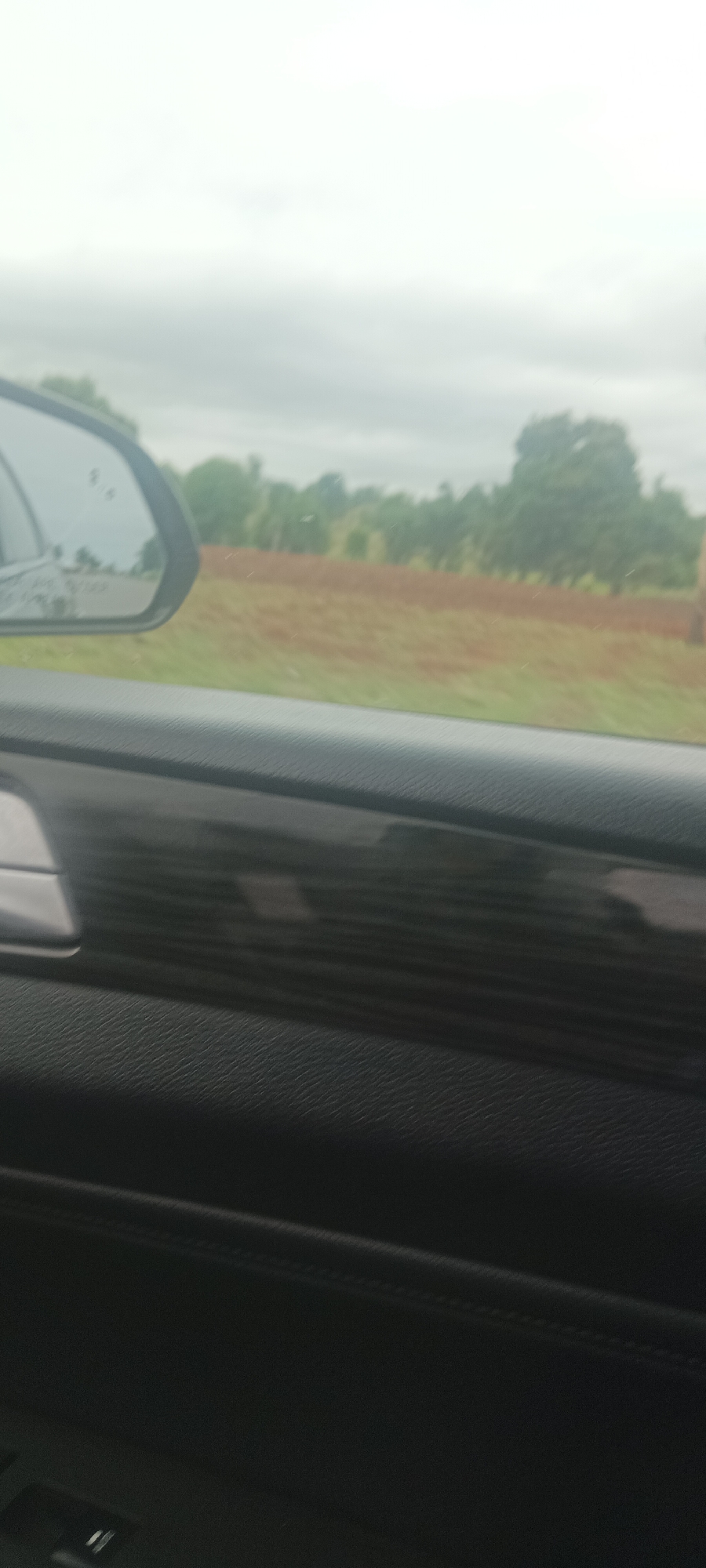 The image shows a view from inside a car, looking out towards a rural landscape. The car's side mirror is visible in the upper left of the frame, reflecting a blurry view of the road and surrounding trees. Beyond the car's interior, a field or open space with reddish-brown soil is visible. A line of trees is present along the horizon. The sky is overcast. The car's dashboard and door panel are in the foreground, with a black textured surface and a glossy wood-grain trim. The location is Kadarko, Nigeria.
