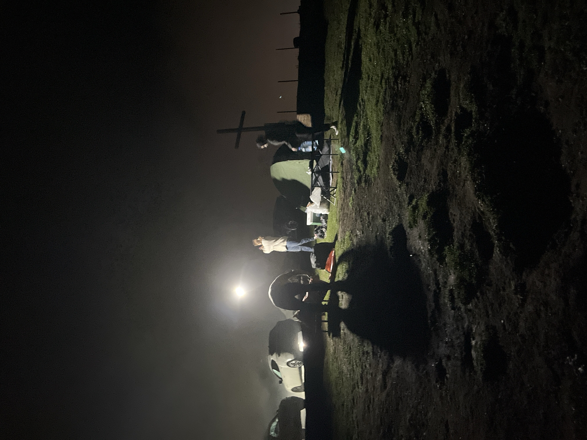 In the city of Gomismta, Georgia, at night, several individuals are engaged in camping activities. The scene is illuminated by a bright light source, possibly a portable lamp, casting strong shadows. A white car is parked to the left, partially obscured by fog or mist. In the center of the image, a person is bent over what appears to be a portable stove or grill. To the right, a green tent is set up, and a person is carrying a large jug of water. Further to the right, a wooden cross stands against the dark sky. The ground is covered in grass and dirt, with some evidence of rough terrain. The overall atmosphere is one of a nighttime outdoor gathering or campsite, with the natural elements of mist and darkness contributing to the scene.