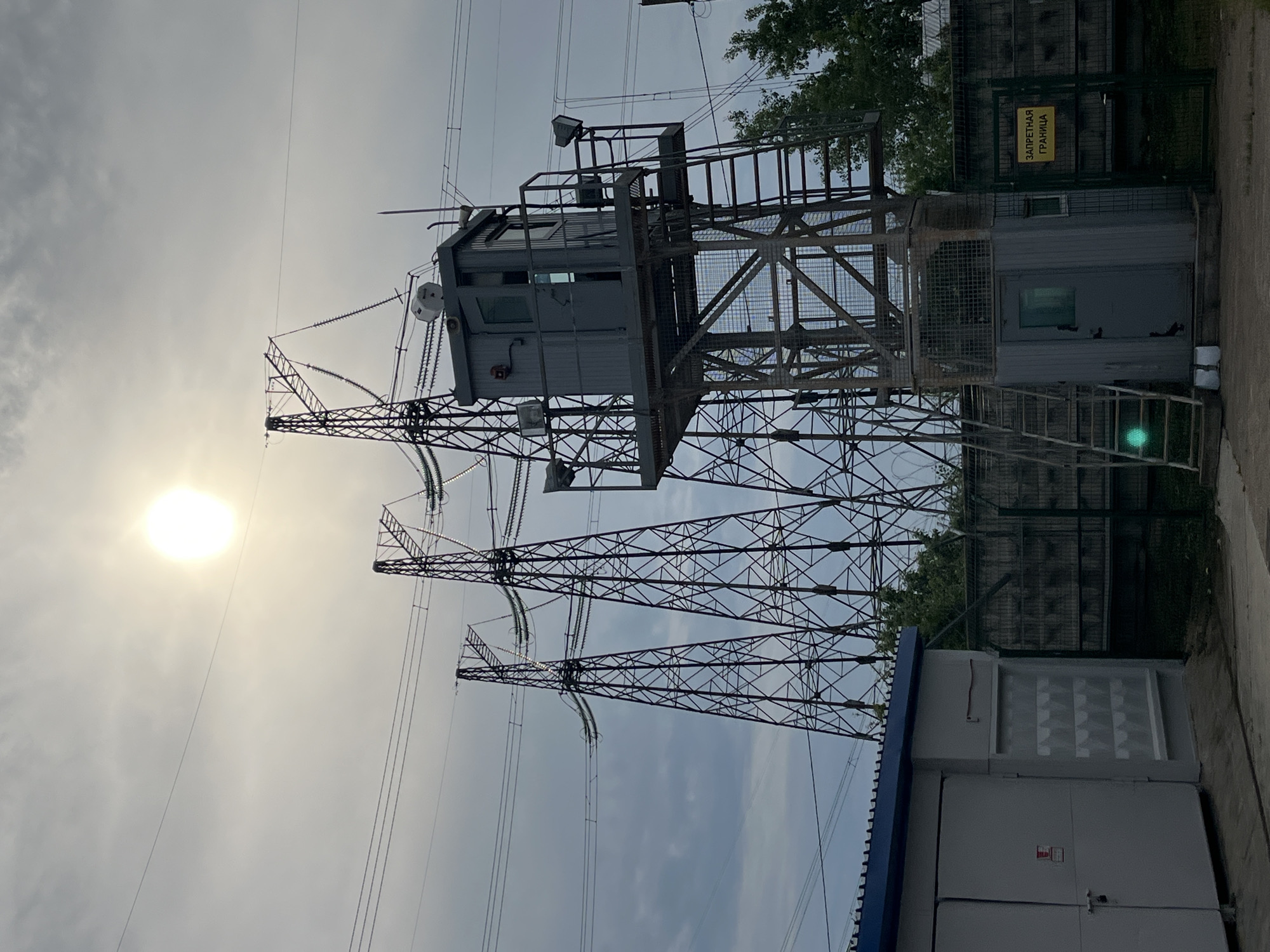 The image displays an industrial outdoor scene featuring high-voltage power transmission infrastructure in Chaikovsky, Russia, under a bright, hazy sky. No people are visible.
Dominating the central and left portions of the frame are several towering, lattice-steel electricity pylons, supporting numerous thick power lines that stretch across the sky. These pylons feature multiple insulator strings and complex bracing.
On the right side of the image, an elevated, enclosed metallic structure resembling a guard booth or control room is mounted on a separate support frame or an extension of one of the pylons. This booth has windows, a small satellite dish or sensor, and is accessible via a metal staircase. Below and to its right, a grey, modular building or container with a door and a window is situated behind a fence. Further to the right, in the foreground, is a larger, rectangular utility building or transformer station with a blue slatted roof and grey panels.
A dark mesh or chain-link fence runs along the right side of the frame, enclosing the area. Behind the fence and some of the structures, green foliage from trees is visible. The sun appears as a bright, circular disc in the upper left part of the hazy sky, suggesting daytime, possibly late morning or afternoon.
A yellow sign is clearly visible on the fence near the modular building, bearing the Russian text "ЗАПРЕТНАЯ ГРАНИЦА", which translates to "FORBIDDEN BOUNDARY" or "PROHIBITED BORDER," indicating restricted access to the site. A small, red warning sign is present on the door of the utility building in the foreground, but its text is illegible. The overall scene depicts a functional, secured electrical power transmission site.