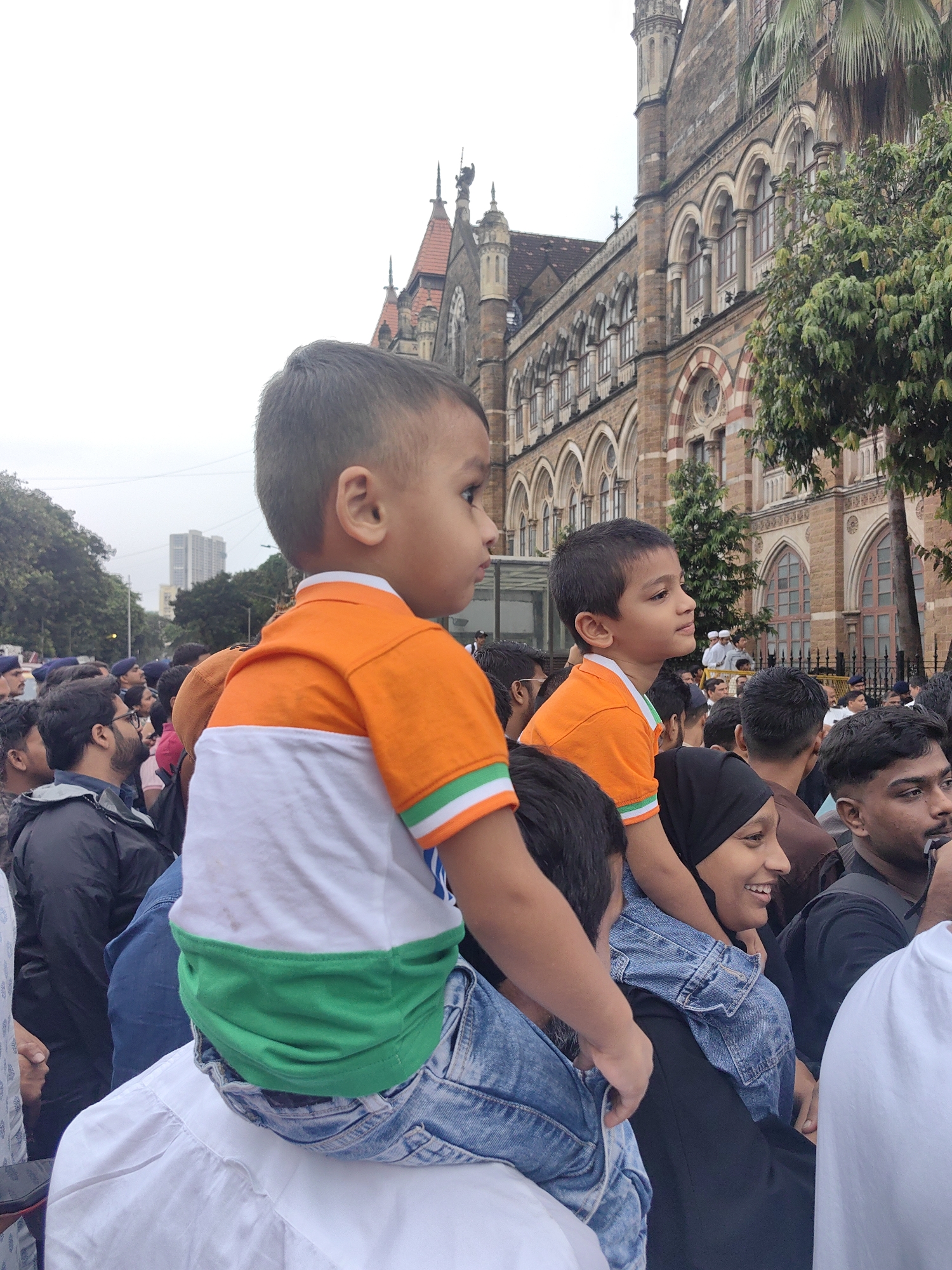 A group of people are gathered outdoors, with many wearing clothing in the colors of the Indian flag (saffron, white, and green). In the foreground, a young boy is being carried on someone's shoulders, wearing an orange shirt with a white and green band across the chest, resembling the Indian flag. Another boy, also wearing a similar shirt, is visible on the shoulders of another person behind him.
The background features a large, ornate, historic building with multiple arched windows and intricate stone detailing, consistent with Victorian Gothic architecture. This building is identifiable as the Chhatrapati Shivaji Maharaj Terminus (CSMT) in Mumbai, India, formerly known as Victoria Terminus. Trees and a street are also visible, suggesting an urban environment. The sky is overcast.
The overall scene depicts a public gathering, possibly a patriotic event or celebration, with families and individuals present. The focus on the children in flag-colored attire highlights a sense of national pride.