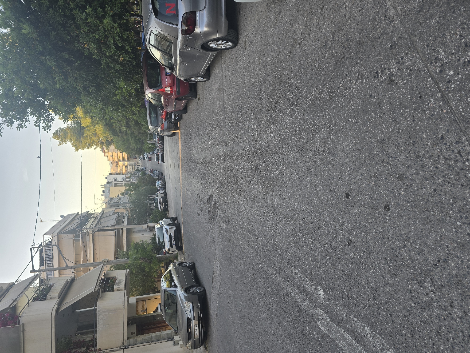 This rotated image captures a residential street, likely in Nea Ionia, Greece, under a clear, bright sky suggesting late afternoon or early evening. The scene is dominated by a narrow asphalt road flanked by parked cars and multi-story apartment buildings.
On the left side of the street, a continuous line of cars is parked. Prominently in the foreground is a dark grey Peugeot sedan with the license plate "MTT 9181". Behind it is a lighter grey hatchback, possibly also a Peugeot, distinguishable by "PEUGEOT" text on its tailgate and a red 'N' decal on its rear window. Further down the row, a red Citroën, with "CITROEN" visible on its back, is also parked. The cars are varied in model and color, suggesting a typical urban parking scenario.
Opposite, on the right side of the street, another row of vehicles is parked, including a white car with a partially visible license plate "YXX 0707". The buildings lining both sides of the street are apartment blocks, featuring multiple balconies, some equipped with light-colored retractable awnings. Lush green trees are interspersed along the sidewalks, casting some shadows and adding greenery to the urban landscape. Utility poles and overhead wires are visible against the bright sky. The street itself is made of asphalt, showing some texture and faint road markings. There are no people visible, and no specific event or activity is taking place, presenting a quiet, everyday street scene.