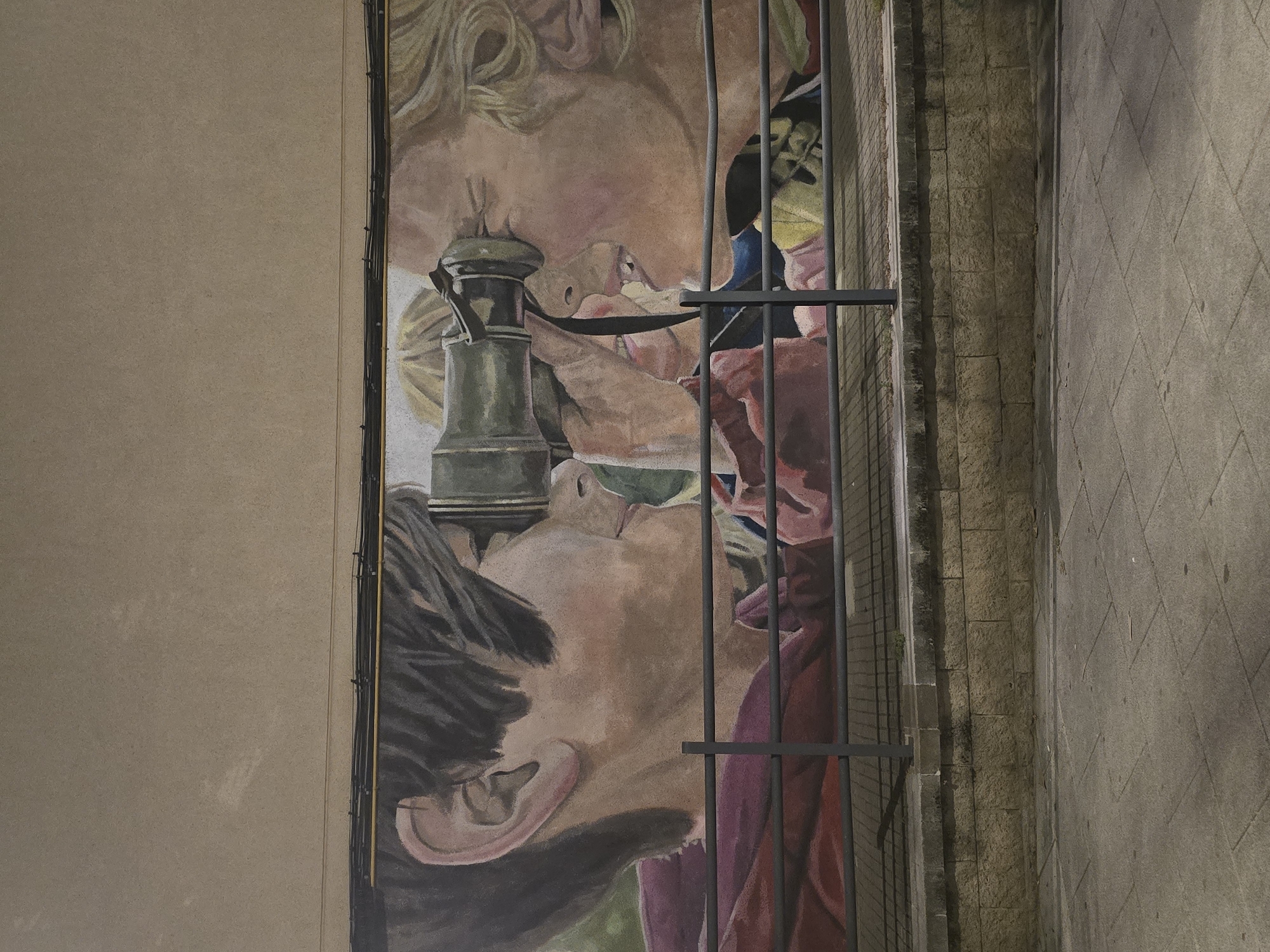 The image displays an outdoor mural on a building wall in Cornellà De Llobregat, Spain. The mural, painted in a realistic style, depicts three young individuals in profile, primarily focusing on two figures interacting with a telescope or spyglass. The leftmost figure, with short dark hair, wears a maroon top and looks into the eyepiece of an antique-style, possibly brass or oxidized green, telescope. The figure on the far right, with blonde, wavy hair, looks into the opposite end of the telescope, with their mouth slightly open. A third partial face is visible between these two, positioned behind the telescope.
A dark grey metal railing spans horizontally across the lower portion of the mural, partially obscuring the artwork. Below the railing, a low wall constructed from light brown, rectangular-cut stone blocks forms a base. In the foreground, a grey paved sidewalk, composed of square tiles, is visible, showing scattered small debris. Above the mural, the building wall is a plain light brown, with multiple bundles of dark electrical wires and a thin yellow conduit running horizontally across it. Shadows from the railing and unseen overhead elements are cast onto the stone wall and sidewalk, indicating artificial light sources in a low-light environment.