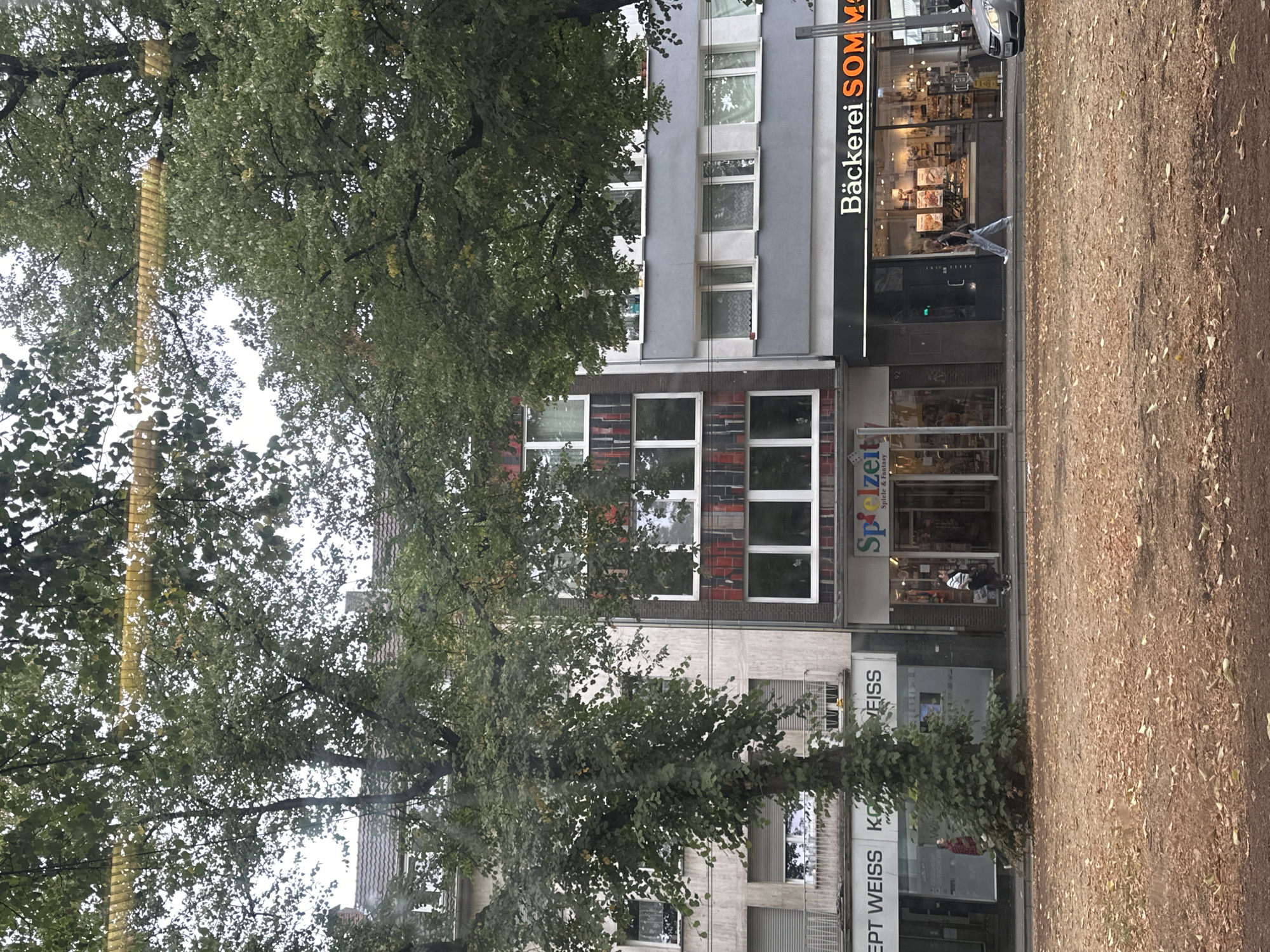 This is a street-level, eye-level, medium shot captured in Krefeld, Germany, focusing on a row of commercial establishments. A large tree with dense green foliage partially obstructs the view of the buildings. The buildings are multi-story structures with modern architectural features.
On the left, a building with a light-colored facade has large windows and awnings. Text is visible on the lower part of this building, including "CEPT WEISS KG" and "VEISS".
Adjacent to it, a building features a vertical section with red and black tiled accents, interspersed with large white-framed windows. Below this, the storefront of "Spielzeit" is visible, indicating a toy or game store, with text reading "Spiele & Fantasy" underneath.
To the right of "Spielzeit," a bakery named "Bäckerei SOMM" is prominent, with a large display window showcasing baked goods. The facade of this establishment is dark with white lettering.
A few individuals are present on the sidewalk. One person, wearing a red and black jacket, is seen near the "CEPT WEISS KG" store. Another individual, dressed in a striped top and jeans, walks past the "Bäckerei SOMM" storefront. A third person, in a white top and blue jeans, walks away from the camera towards the bakery. A silver car is partially visible on the right side of the image, parked or moving.
The foreground consists of a ground surface covered with fallen brown leaves, suggesting an autumn setting. The sky above is overcast and appears light grey. The overall impression is a typical urban street scene with shops and pedestrians.