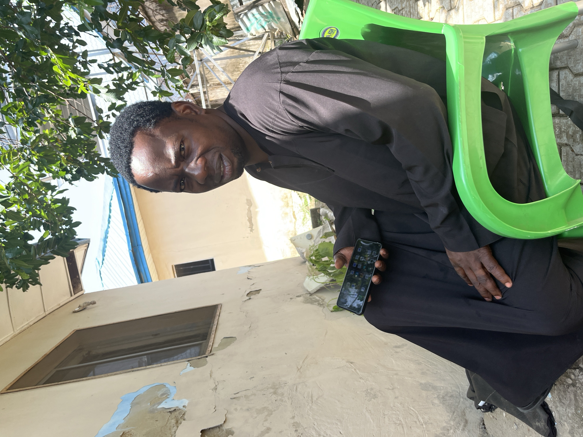 A medium shot captures a man in a black robe sitting on a green plastic chair outdoors. He is looking towards the camera with a serious expression. His right hand holds a smartphone, displaying a grid of app icons. The background shows a cream-colored building with peeling paint and a window, partially obscured by the man's body and the green chair. Lush green foliage hangs from a tree in the upper right portion of the frame. The ground appears to be paved with interlocking bricks. The scene is illuminated by natural daylight.