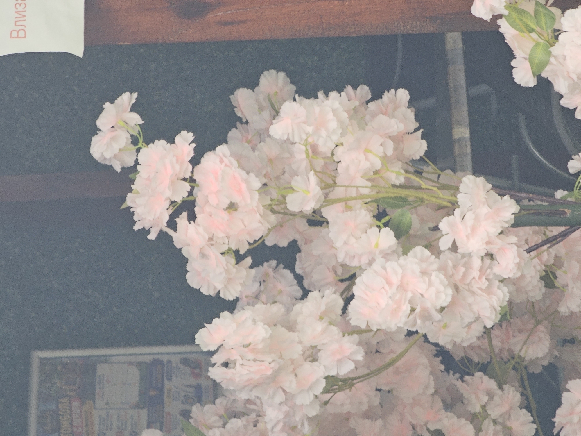 The image shows a close-up of a cluster of artificial light pink cherry blossoms with some green leaves. The blossoms are densely packed, creating a soft, cloud-like effect. In the background, there is a dark, textured wall, and to the left, a blurred poster or sign with Cyrillic text, possibly related to a "TOMOBOLA" (lottery). A portion of a wooden post is visible on the right side of the image. The overall lighting suggests an overcast or indoor setting, as there are no strong shadows. The blossoms appear to be part of a decorative arrangement, likely for an event or establishment.
